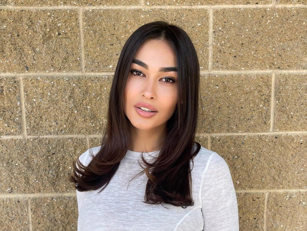 A young woman with long dark hair and light makeup standing in front of a beige brick wall.