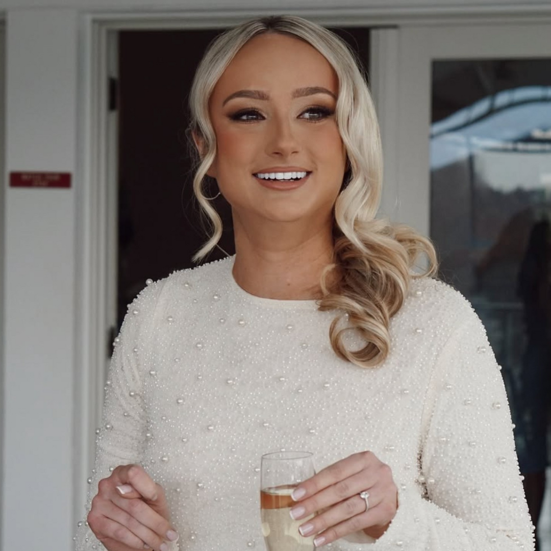 A young woman with long, wavy blonde hair wearing a white lace wedding dress and veil, smiling in an indoor setting.