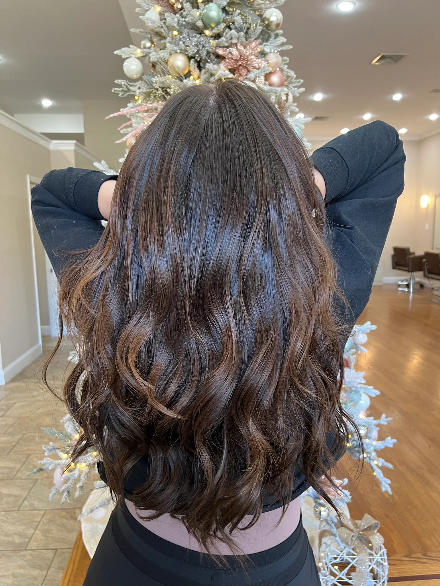 Woman with wavy brown hair standing in front of a decorated Christmas tree in a well-lit room. Her hair has been colored using a balayage technique.