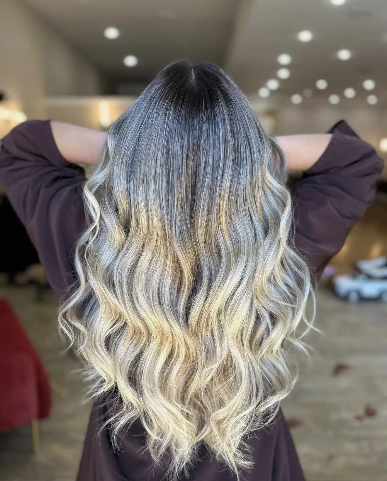 A woman with shoulder-length wavy reddish-brown hair looking down inside a warmly lit interior space. Her hair has been colored using a balayage technique.