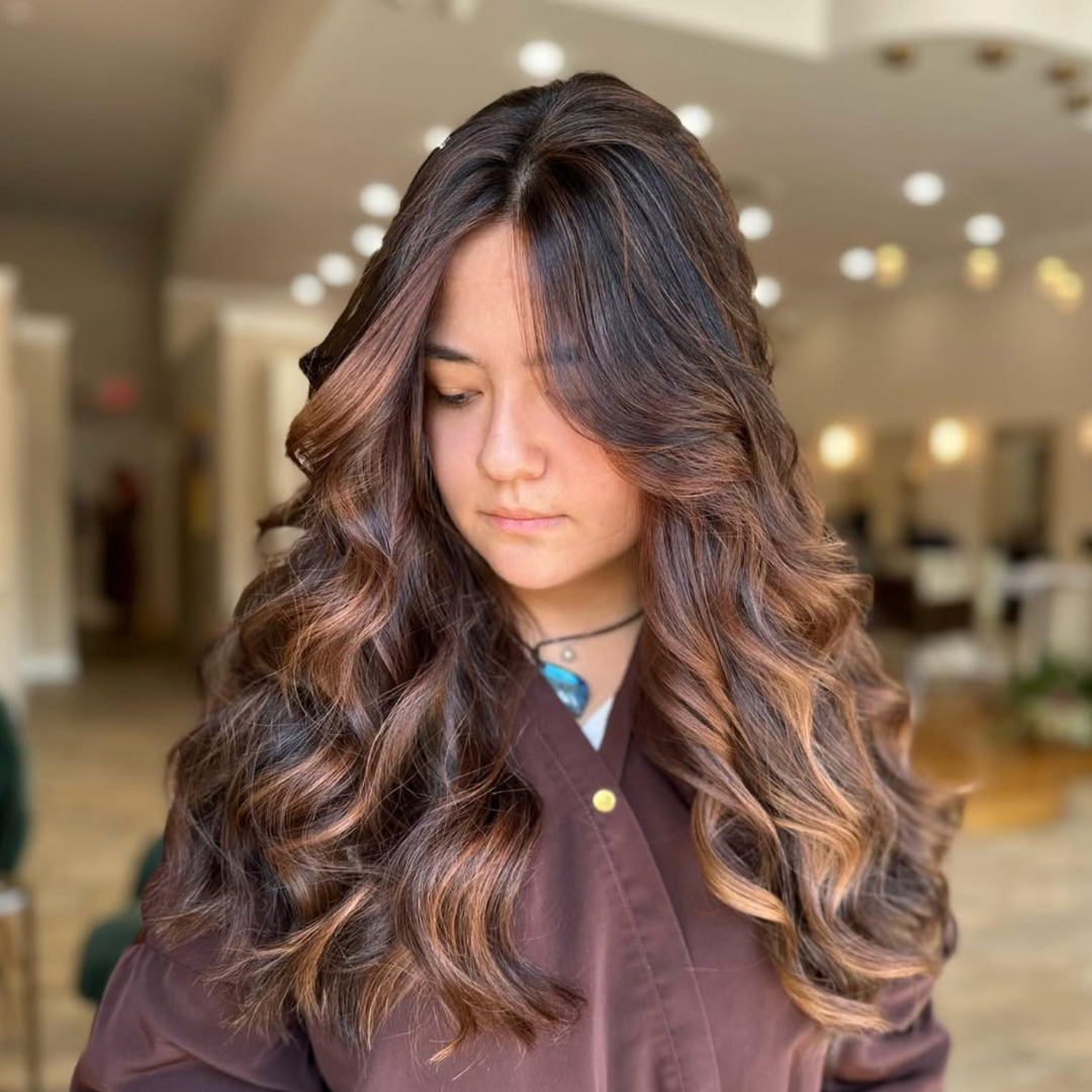 A woman with long, wavy brown hair with lighter highlights, looking down inside a well-lit indoor space.