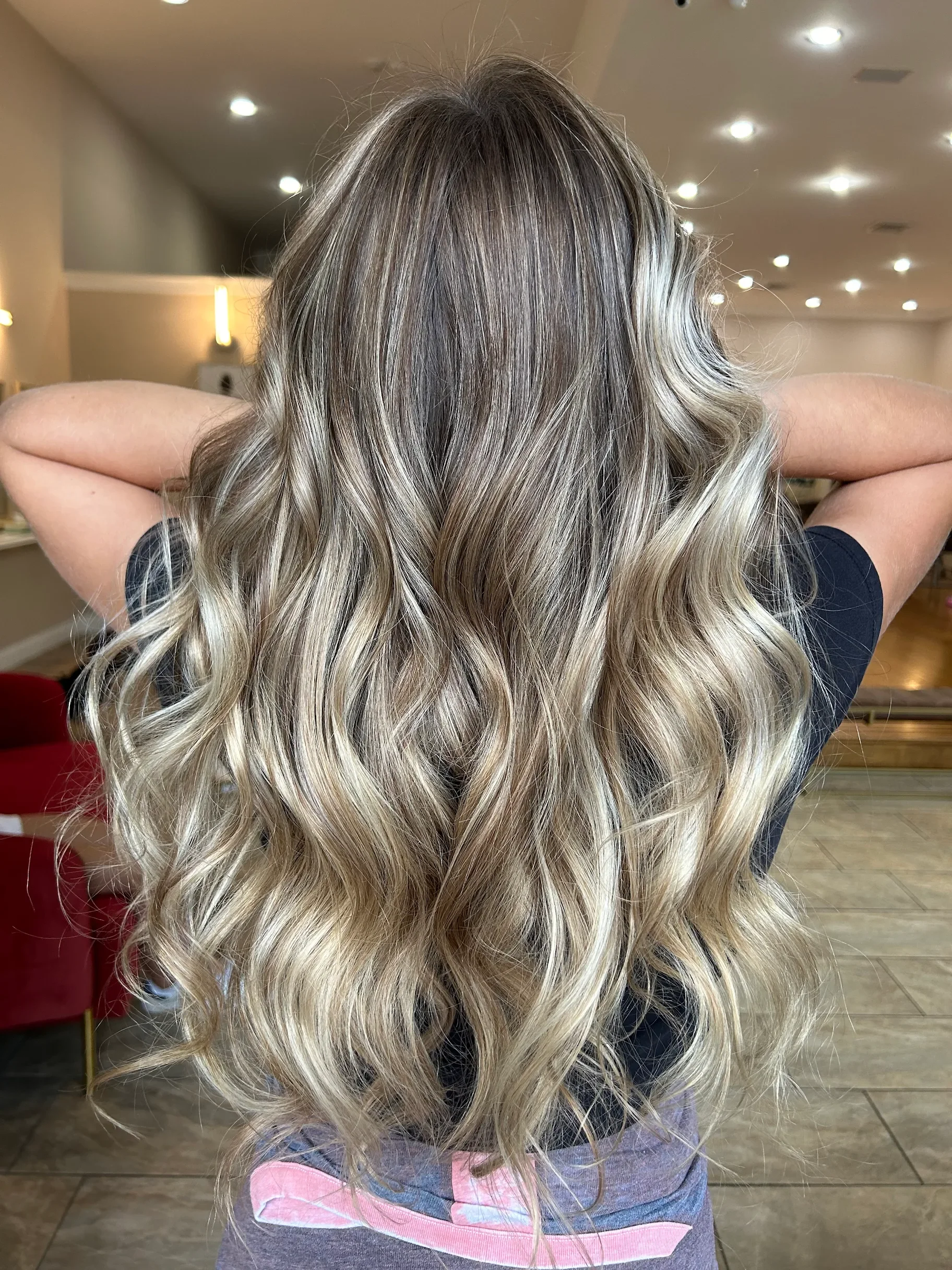 Back view of a woman with long, wavy, blonde hair with darker roots, standing in a salon with warm lighting. She had a brown balayage treatment.