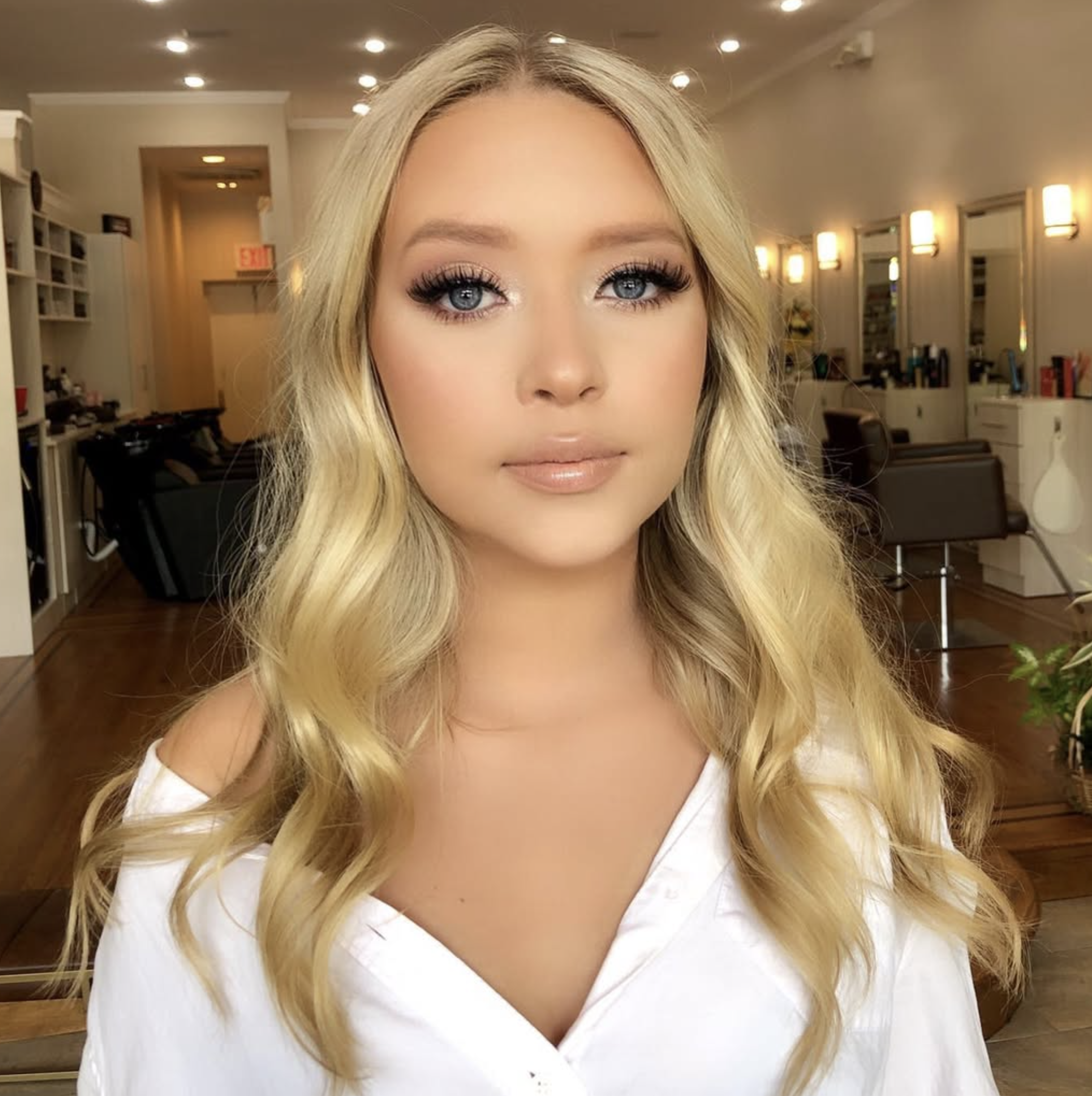 Close-up of a woman's face with blonde hair, blue eyes, and makeup, indoors with warm lighting and bookshelves in the background.