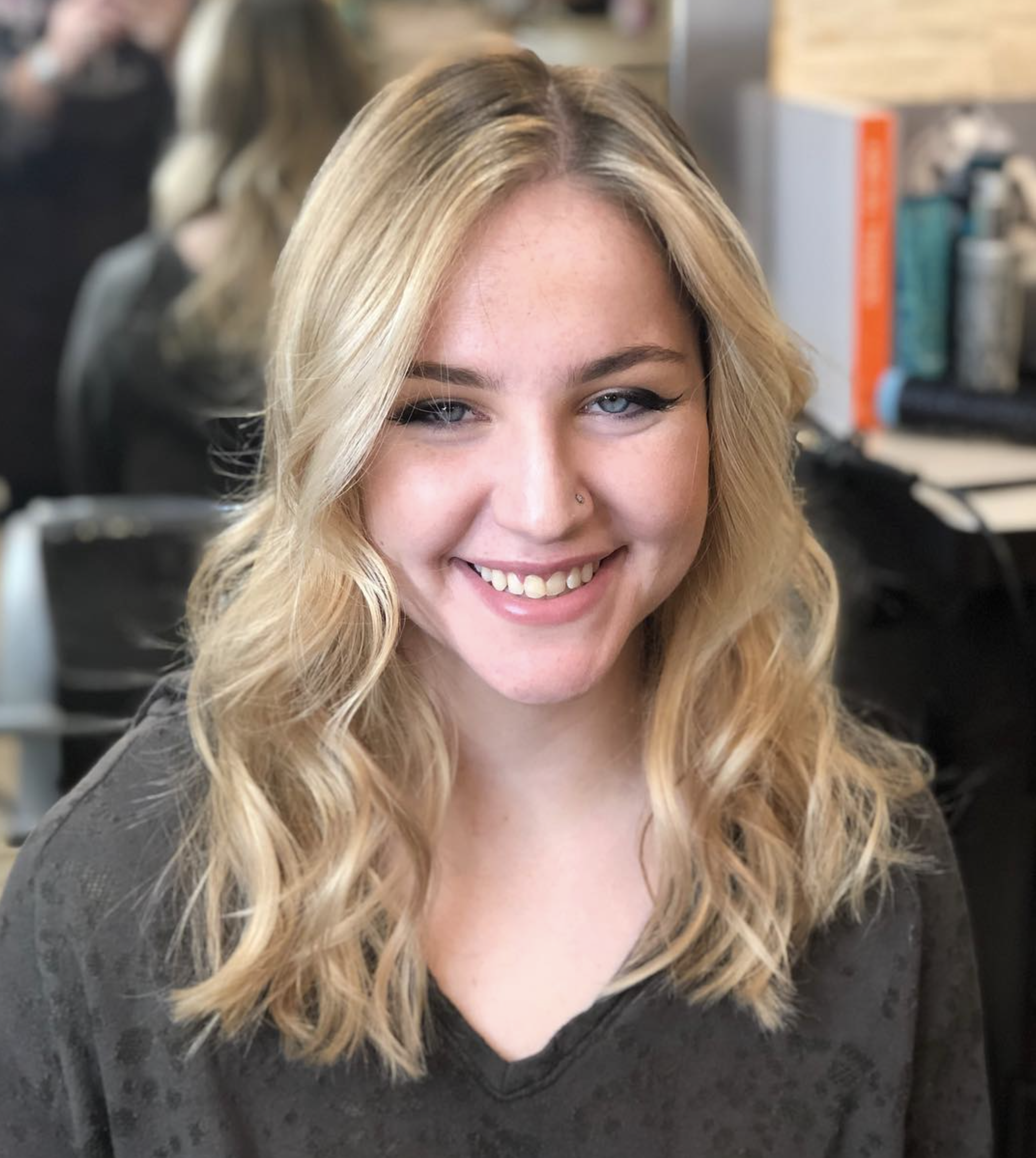 A young woman with blonde, wavy hair and blue eyes smiling at the camera, wearing a black top, in a salon.