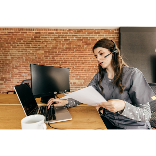 Woman working at a desk with a laptop, monitor, and headset, holding papers in an office with a brick wall background.