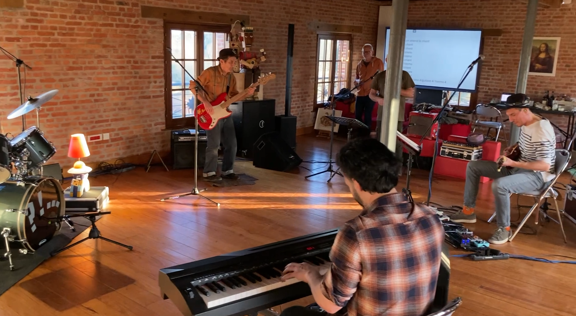Groupe de musiciens en session d'enregistrement dans une pièce avec murs en briques, comprenant un clavier, une guitare, une basse, une batterie, et un synthétiseur, avec un écran blanc en arrière-plan.