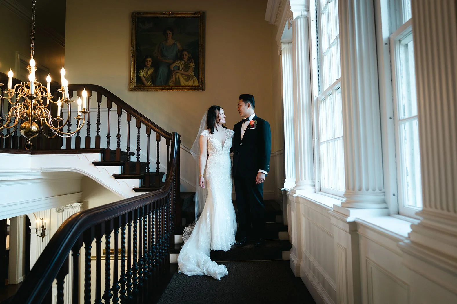 A bride and groom stare at each other on the grand staircase of a mansion