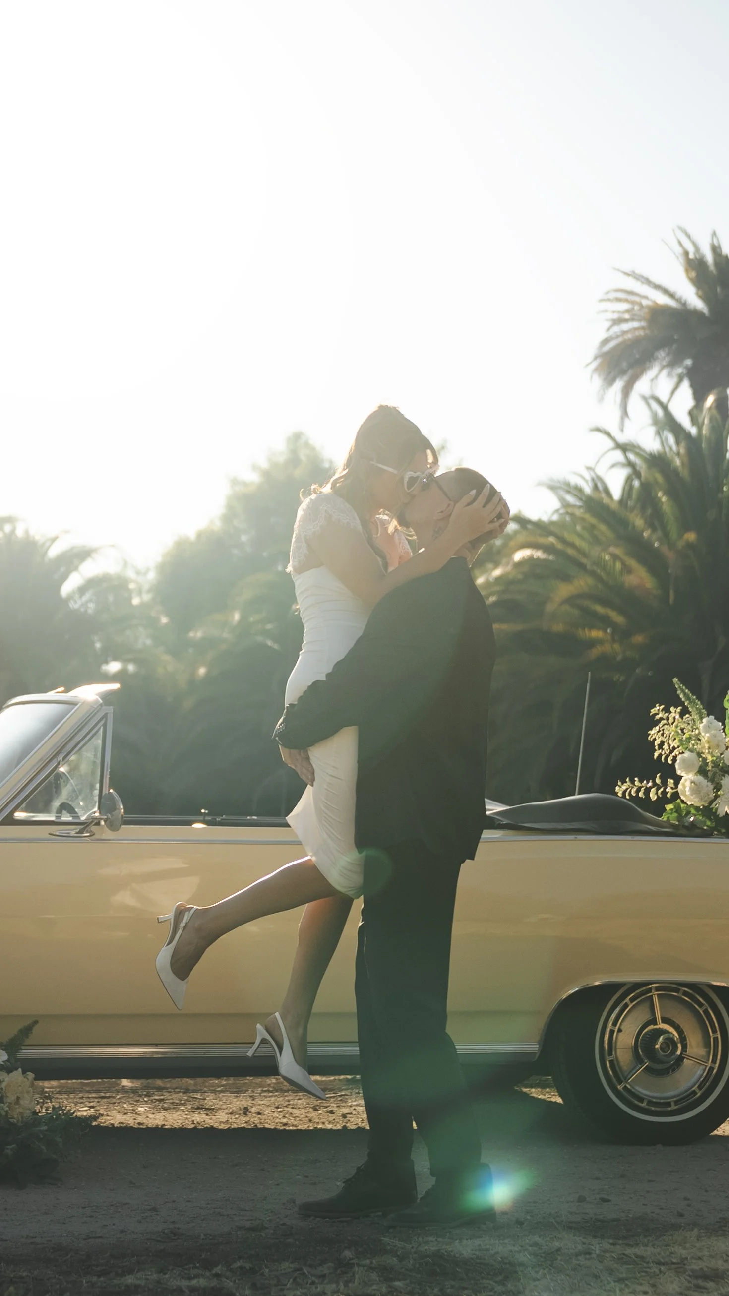 A groom holds a bride in the air as they kiss in front of a yellow convertible.