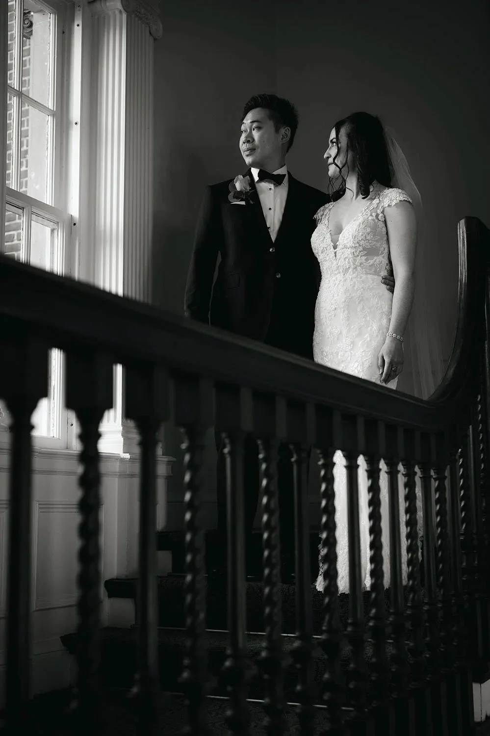 Black and white image of a bride and groom standing on stairs looking out a window