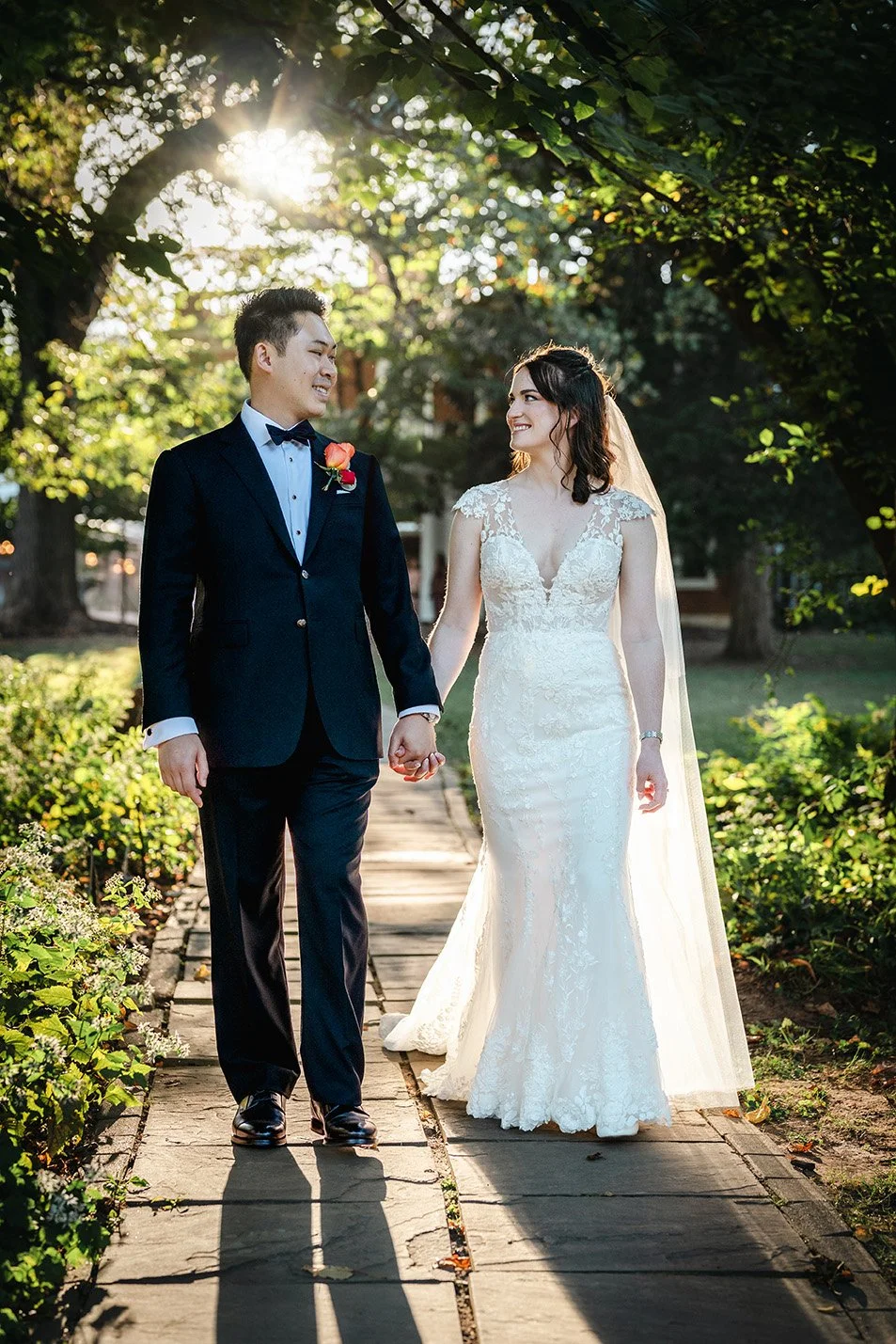 A bride and groom holding hands and smiling at each other on a garden pathway during sunset.