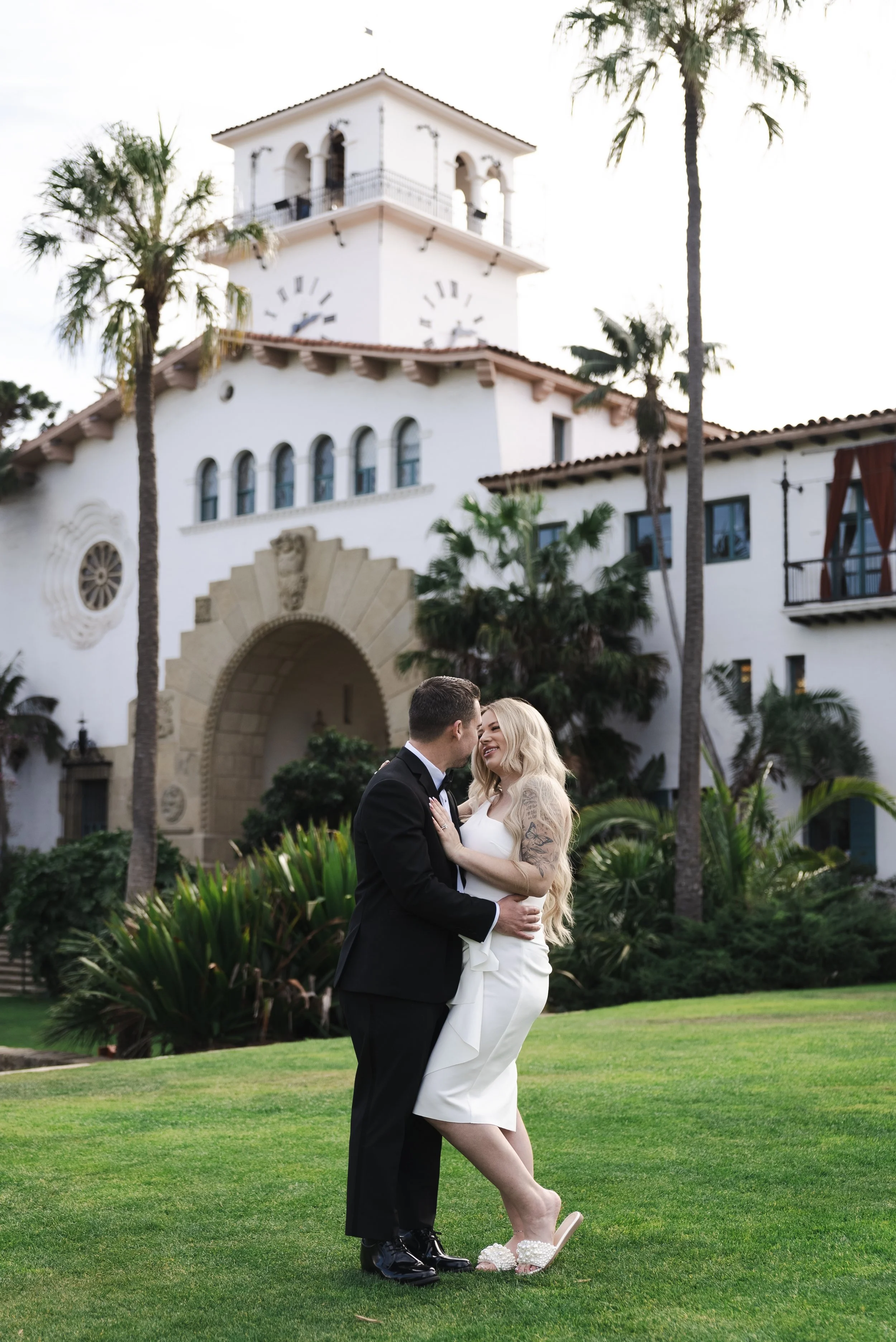 A couple on their wedding day, standing close and smiling at each other, in front of a white building with palm trees, a large arched entrance, and a clock tower, on a grassy lawn.