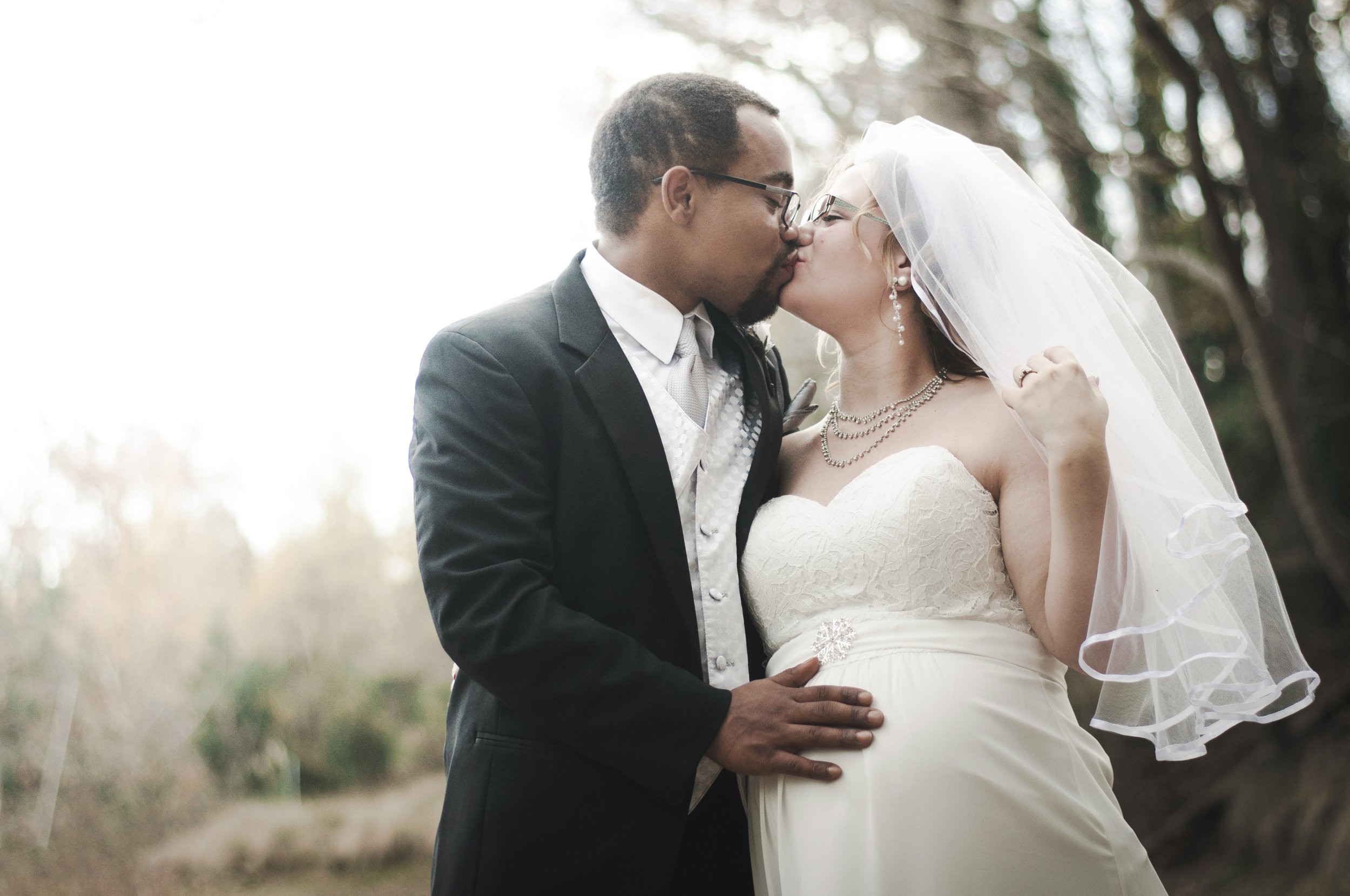 A newlywed couple sharing a kiss outdoors, with the groom in a black tuxedo and the bride in a white strapless wedding gown, wearing a veil and jewelry.
