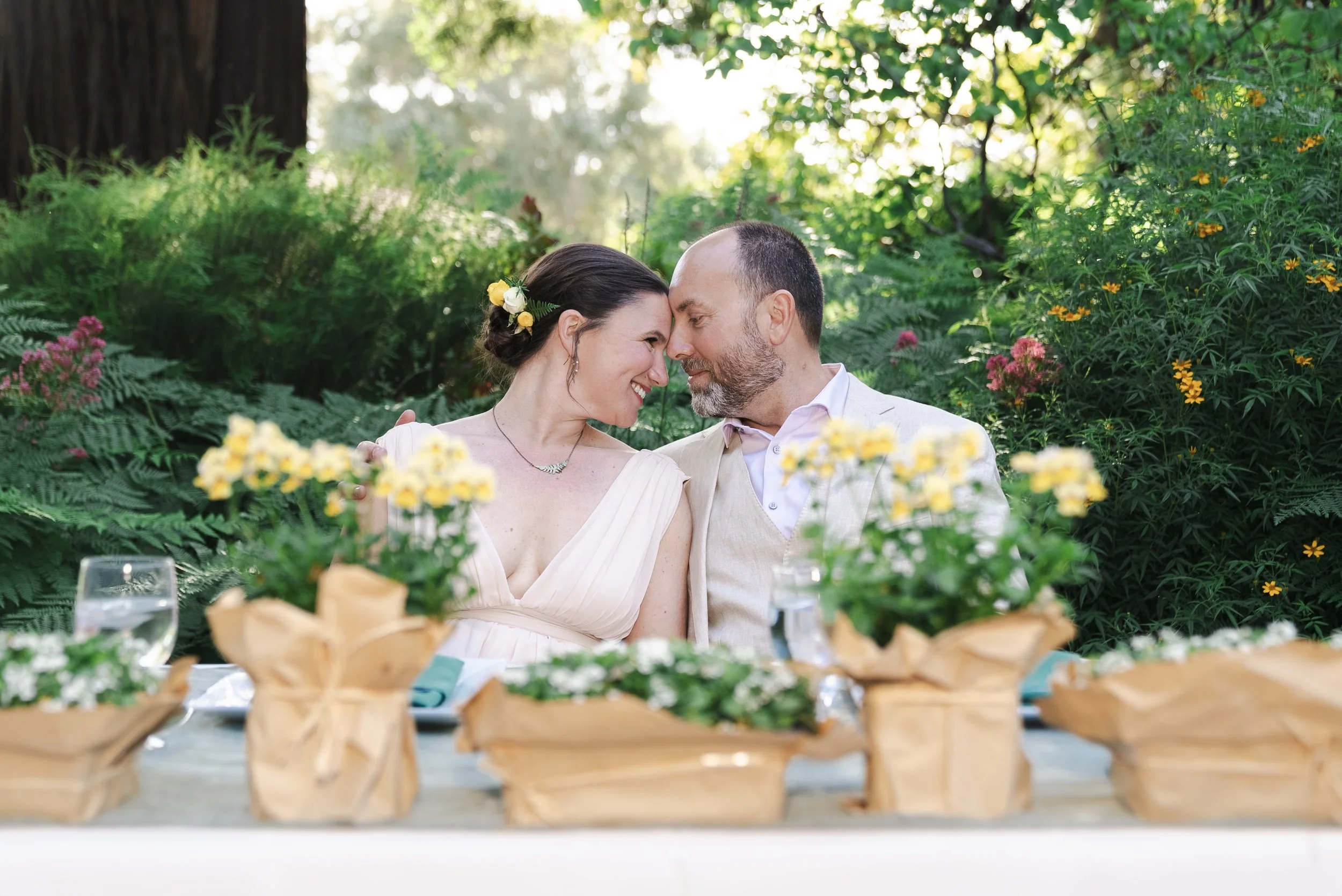 A bride and groom press their heads together at a table in a floral garden