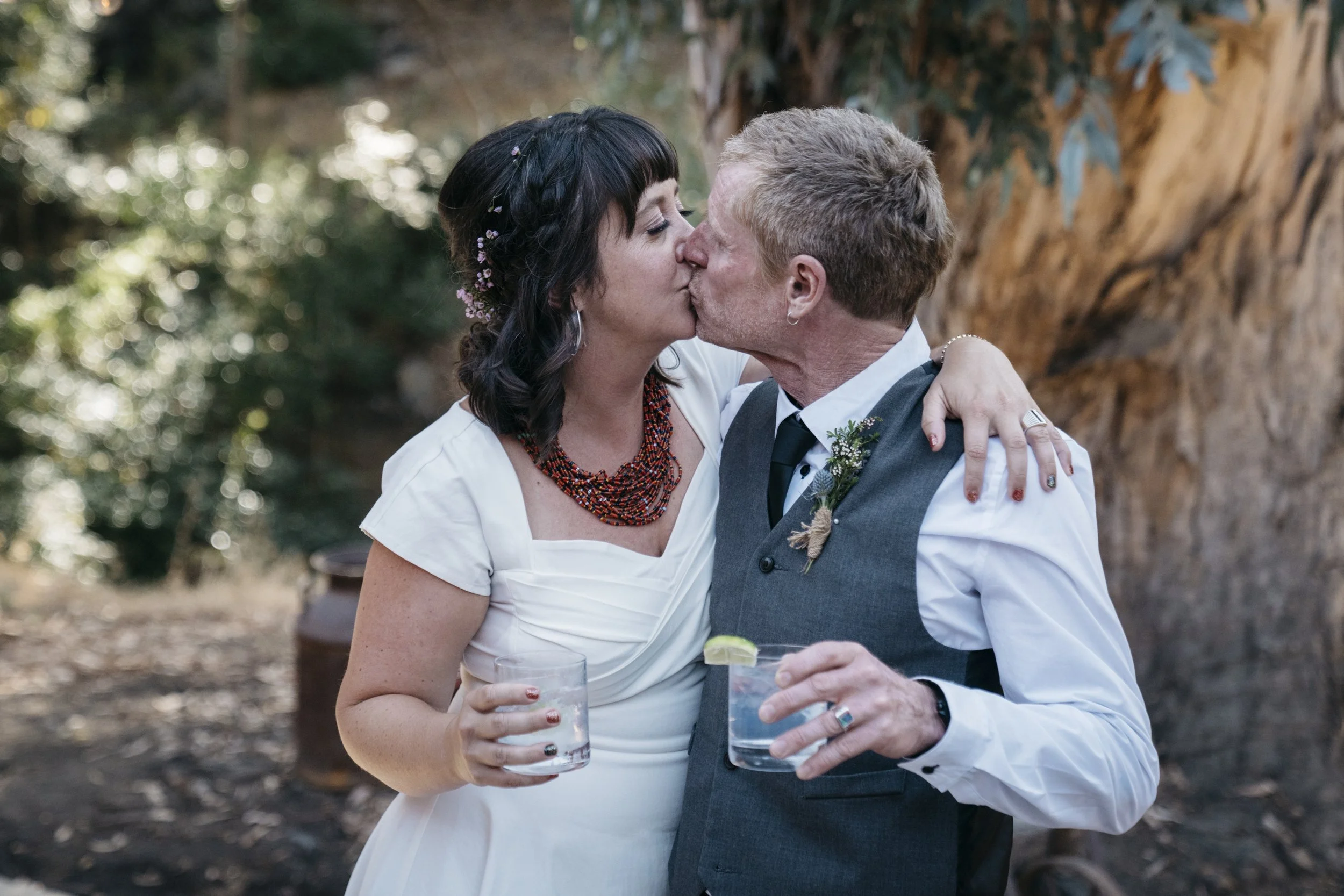A bride and groom holding cocktails kiss each other under a tree