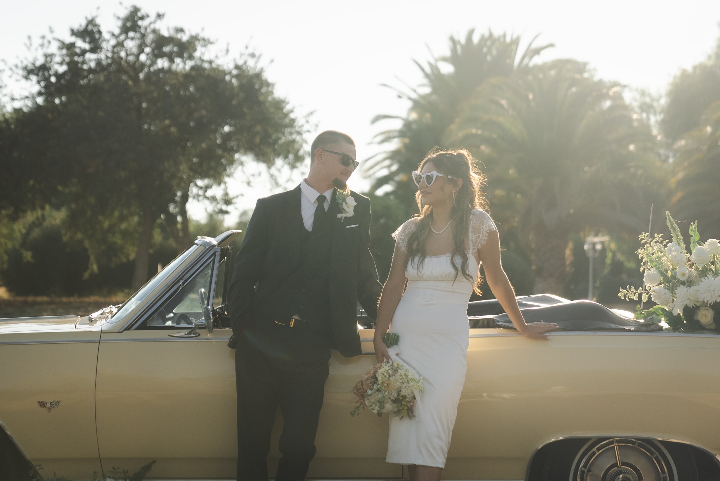 A bride and groom wear sunglasses while leaning against a yellow convertible with palm trees in the background