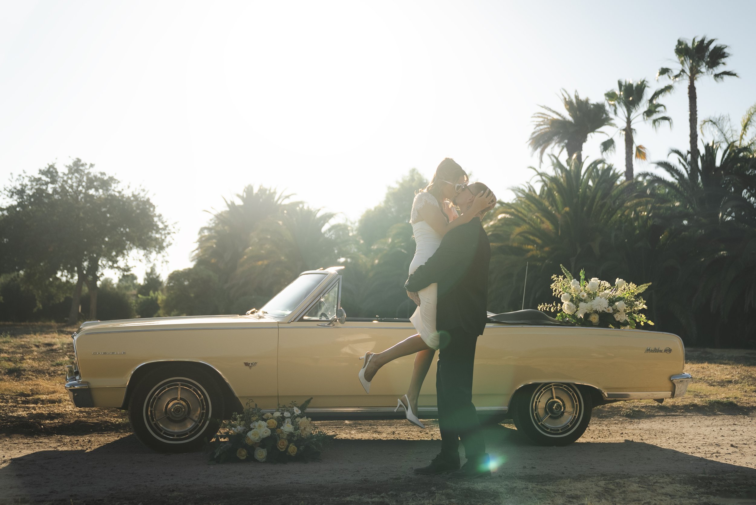 A groom holds a bride in the air next to a yellow convertible car, with palm trees and golden light behind them