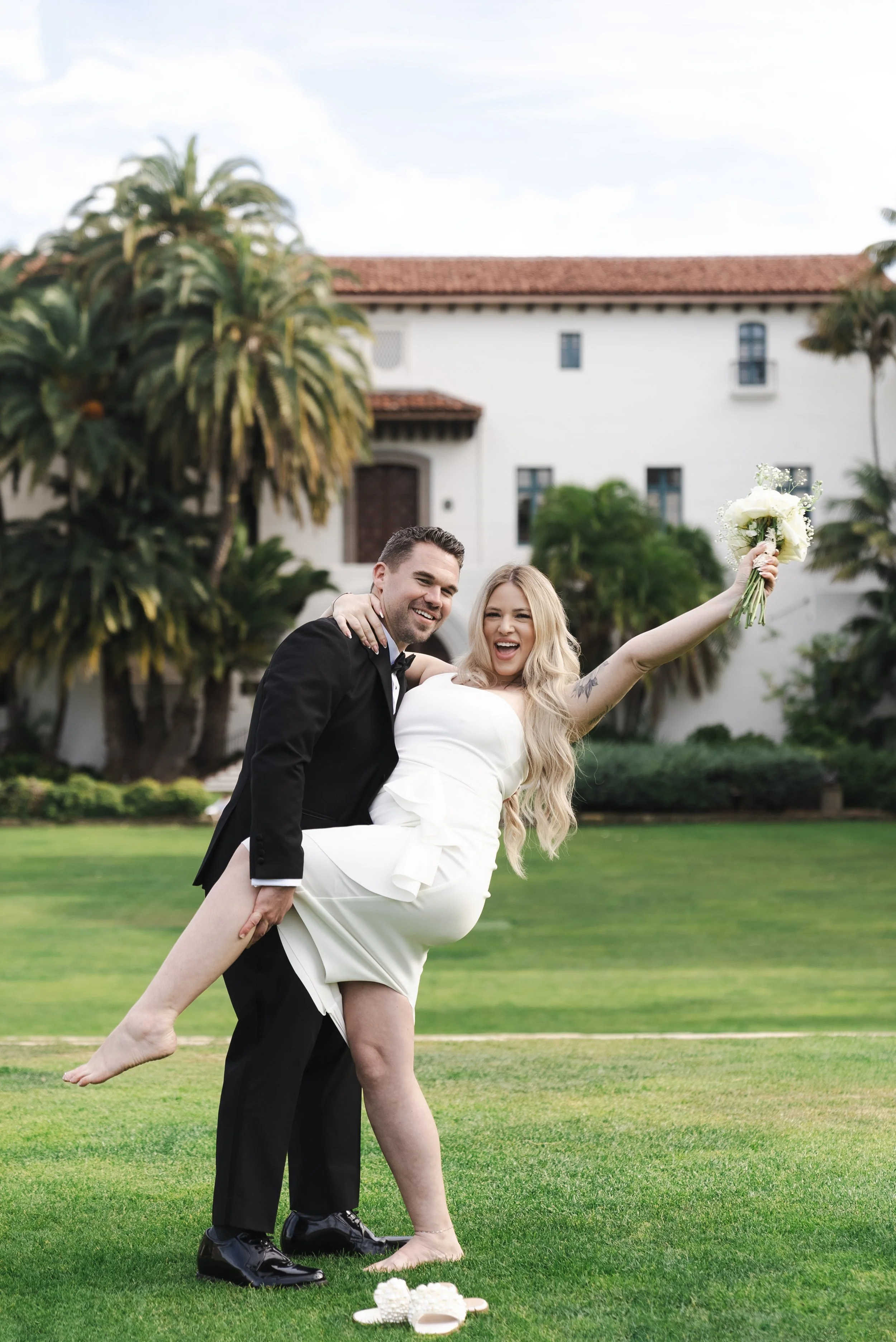 A bride holds flower bouquet in the air as she leans back in the groom's arms in front of the Santa Barbara Courthouse
