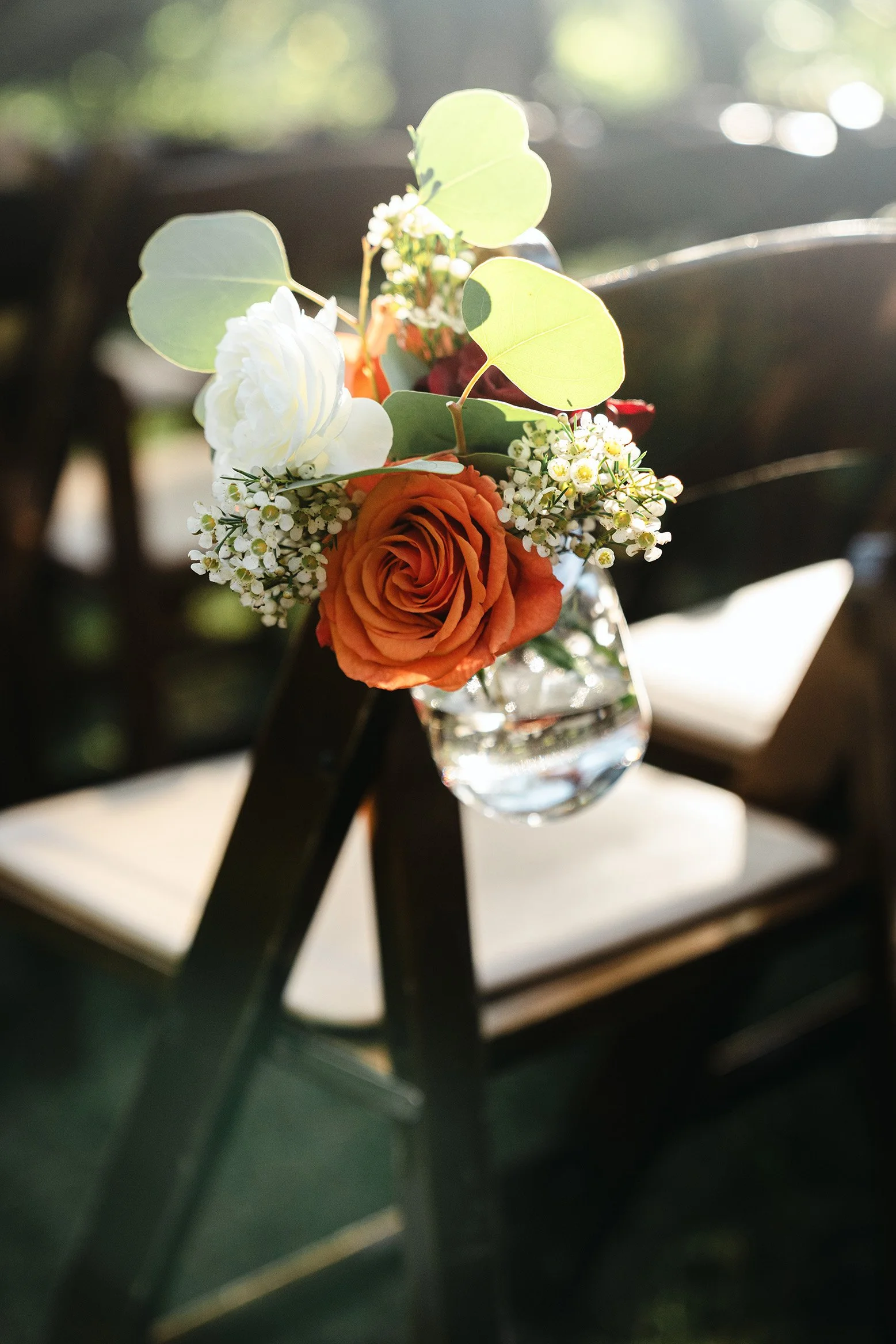 A small floral arrangement with an orange rose, white flowers, and green leaves hanging from the back of a black chair.