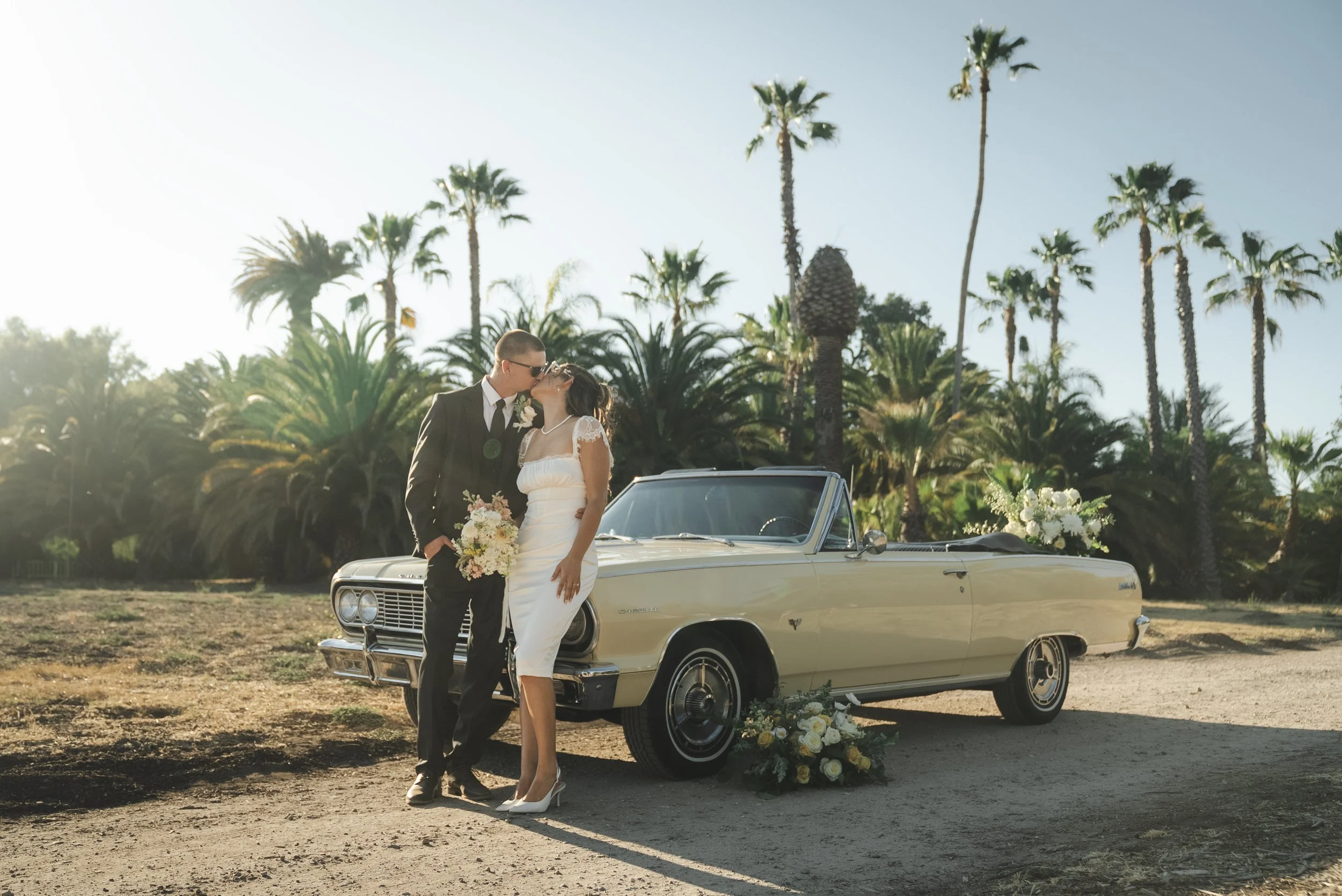 A newlywed couple sharing a kiss in front of a vintage convertible car decorated with flowers, with palm trees in the background during sunset.