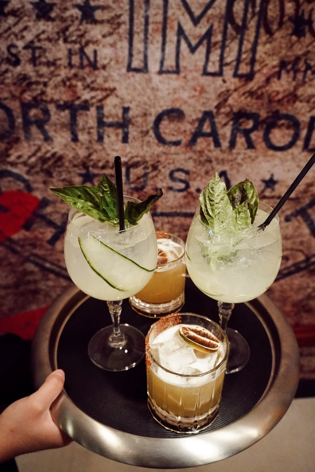 Server carrying a tray of craft cocktails garnished with cucumber, basil, and citrus at The Mecklen Hotel bar in Charlotte, with the hotel’s North Carolina branding mural in the background.