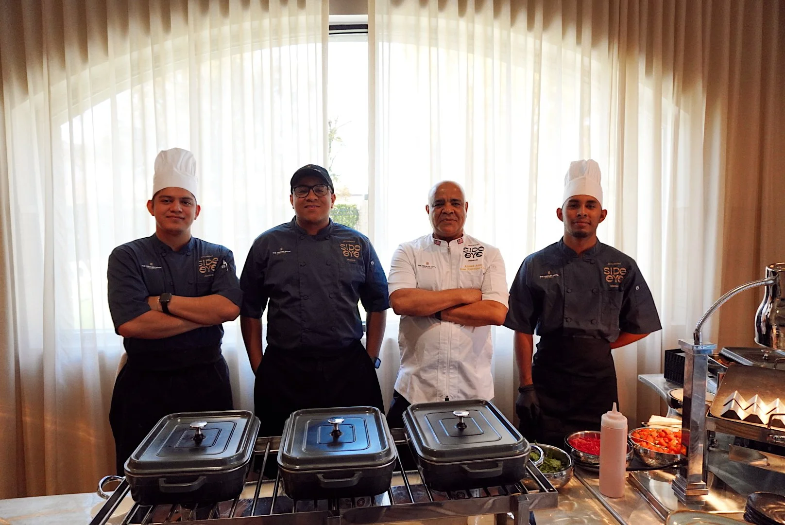 Culinary team from Side Eye at The Mecklen Hotel standing behind a catering station with serving dishes and fresh ingredients prepared for an event.