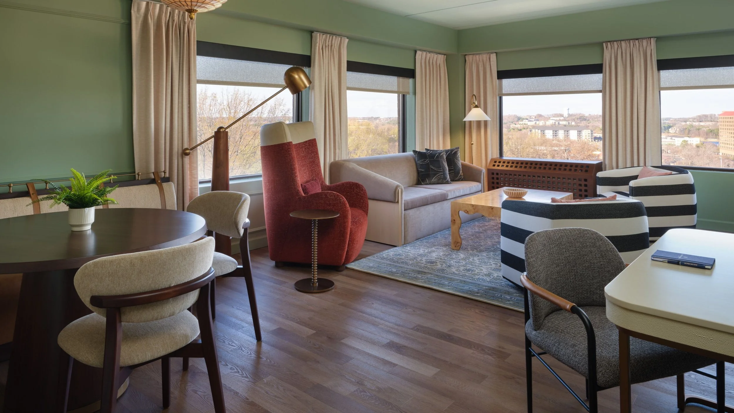 One-bedroom suite living area at The Mecklen Hotel in Charlotte featuring a lounge seating area, dining table, modern furnishings, and large corner windows with city views.