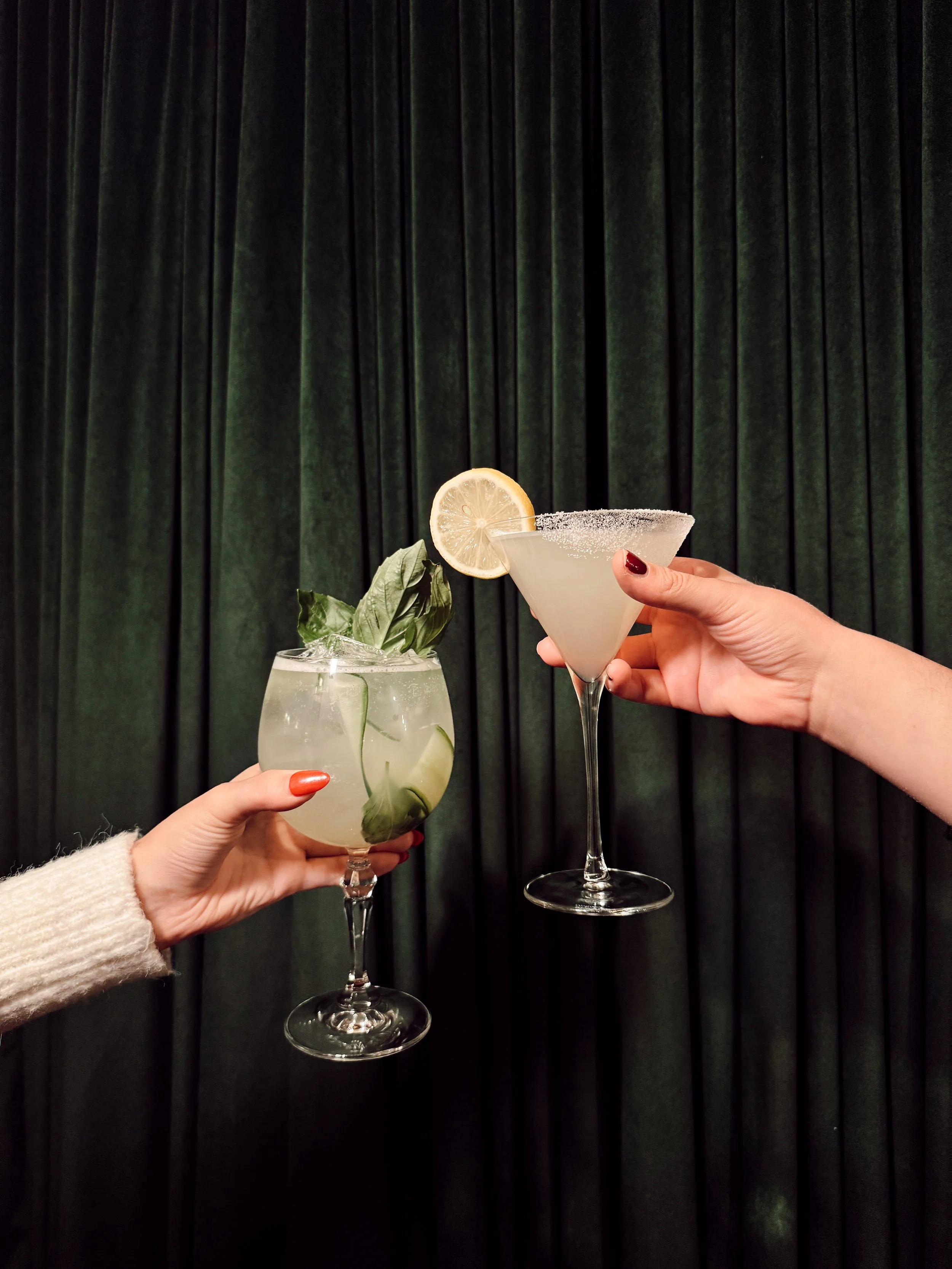 Two hands holding craft cocktails—a basil garnish drink and a lemon-garnished martini—against a dark green curtain backdrop at Side Eye lounge at The Mecklen Hotel.