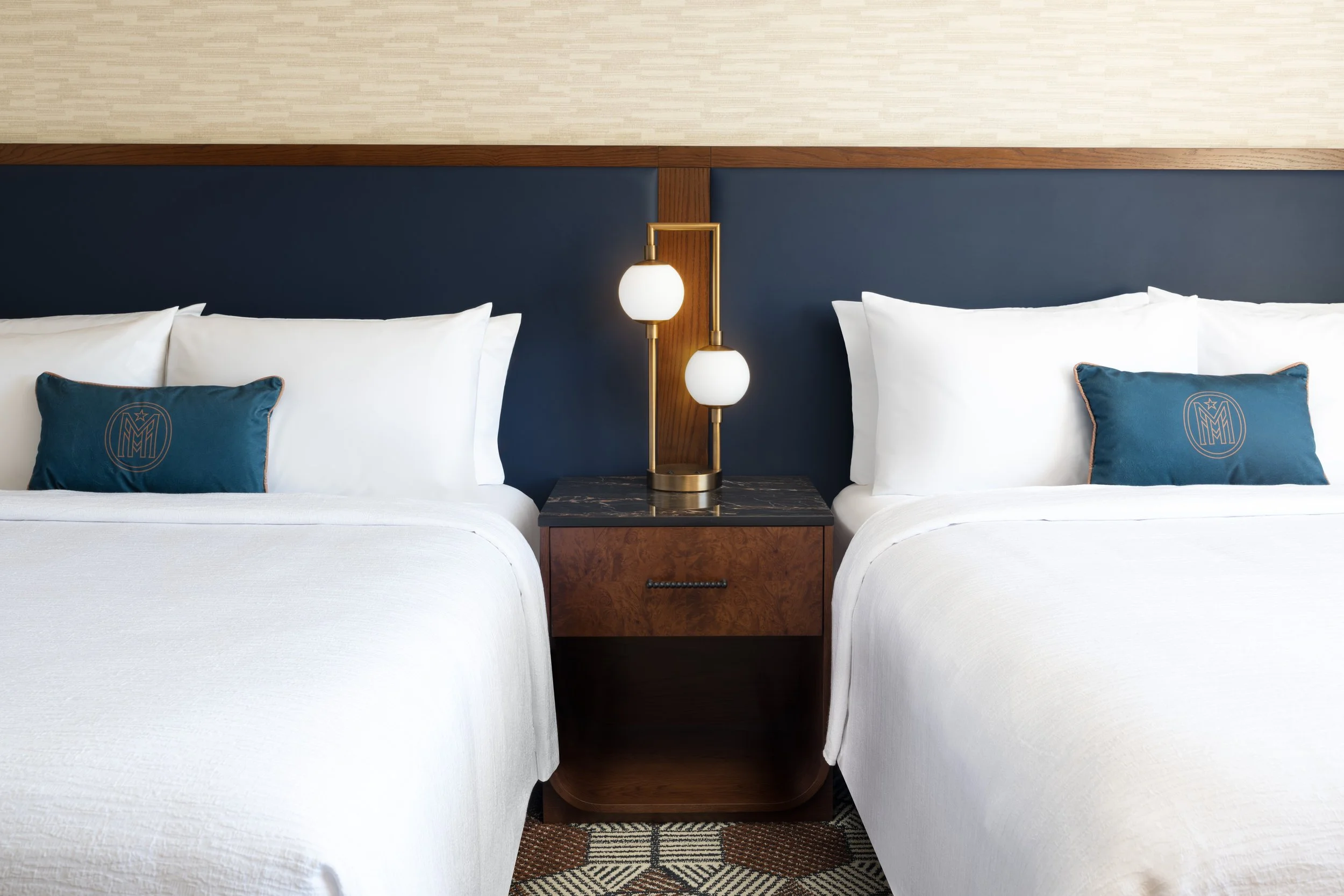 Two queen beds in a guest room at The Mecklen Hotel in Charlotte featuring plush white bedding, branded accent pillows, modern bedside lighting, and wood nightstand.
