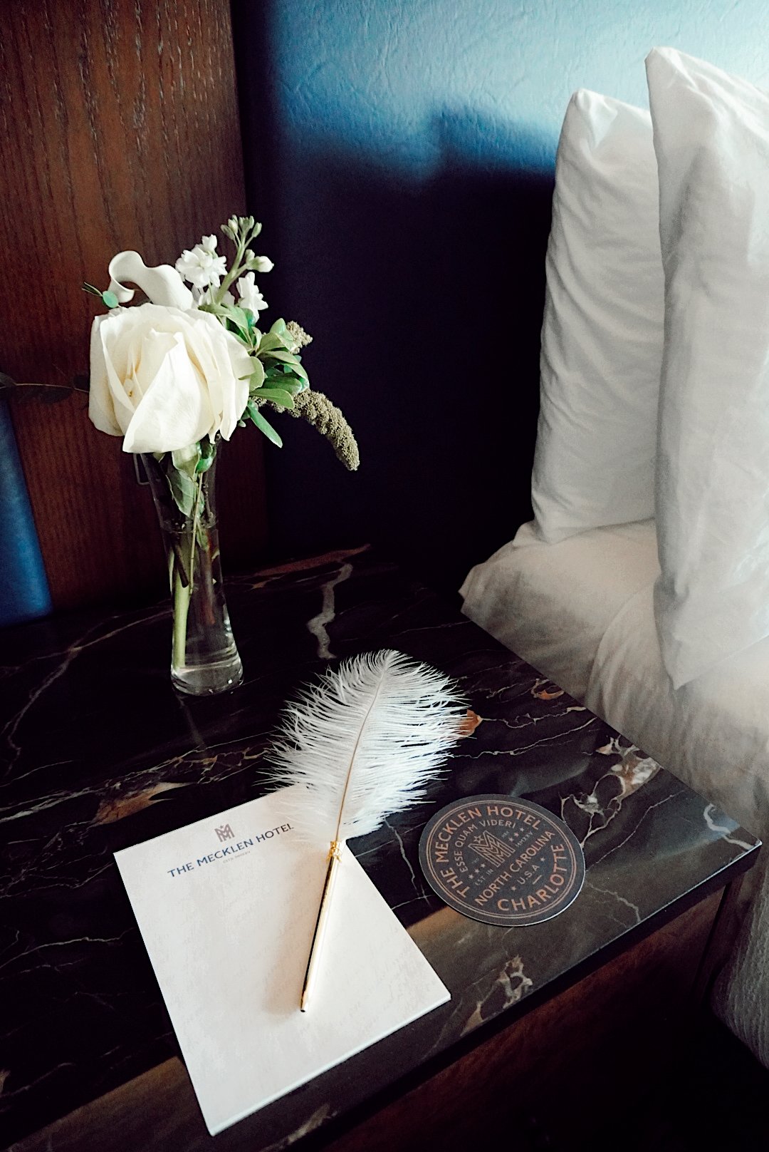 Elegant bedside vignette at The Mecklen Hotel featuring a marble nightstand with a white rose arrangement in a glass vase, a feather quill pen resting on branded stationery, and plush white pillows against a deep blue wall.