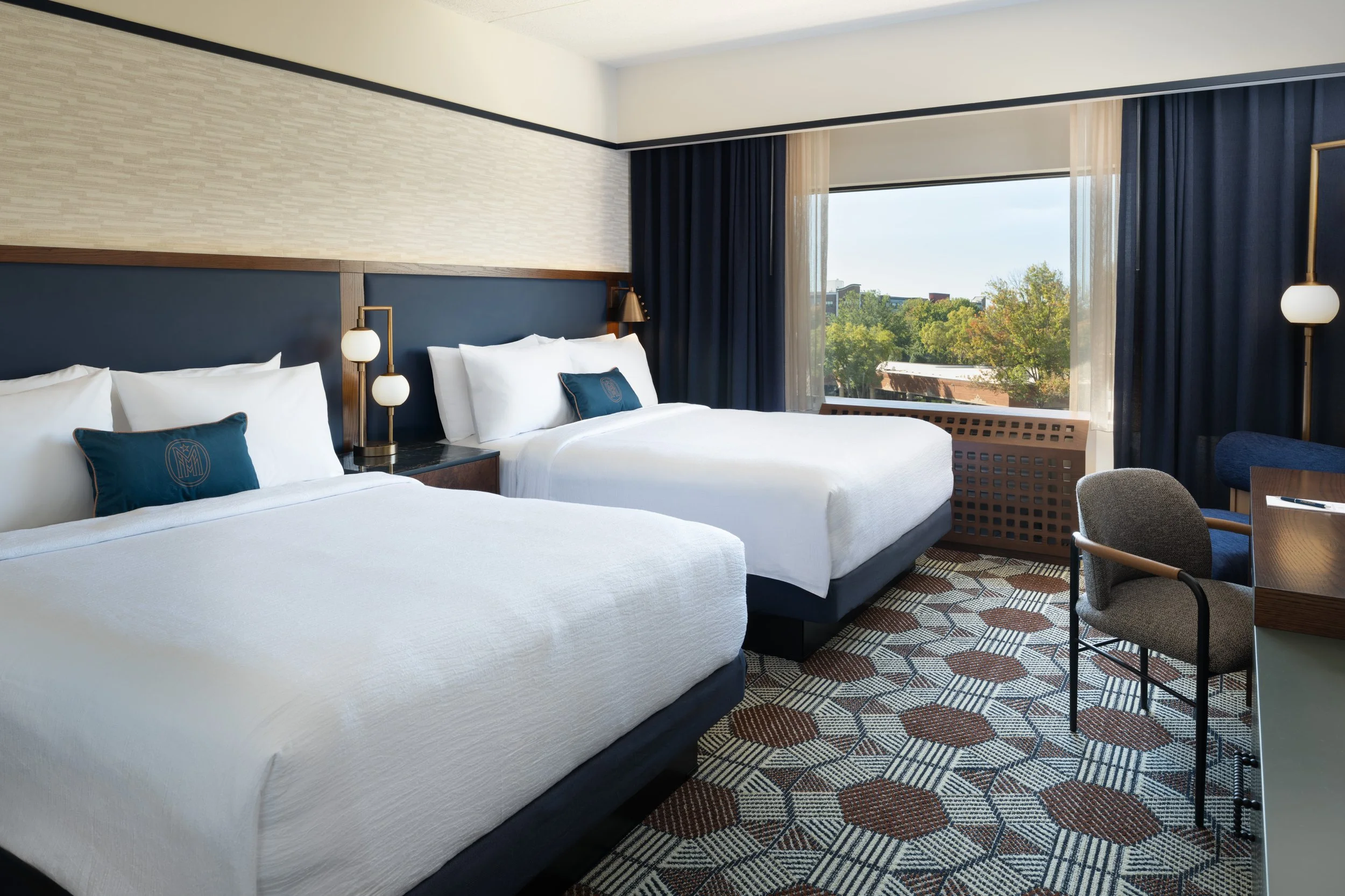 Double queen guest room at The Mecklen Hotel in Charlotte featuring two plush beds, modern lighting, patterned flooring, and large window views with natural light.