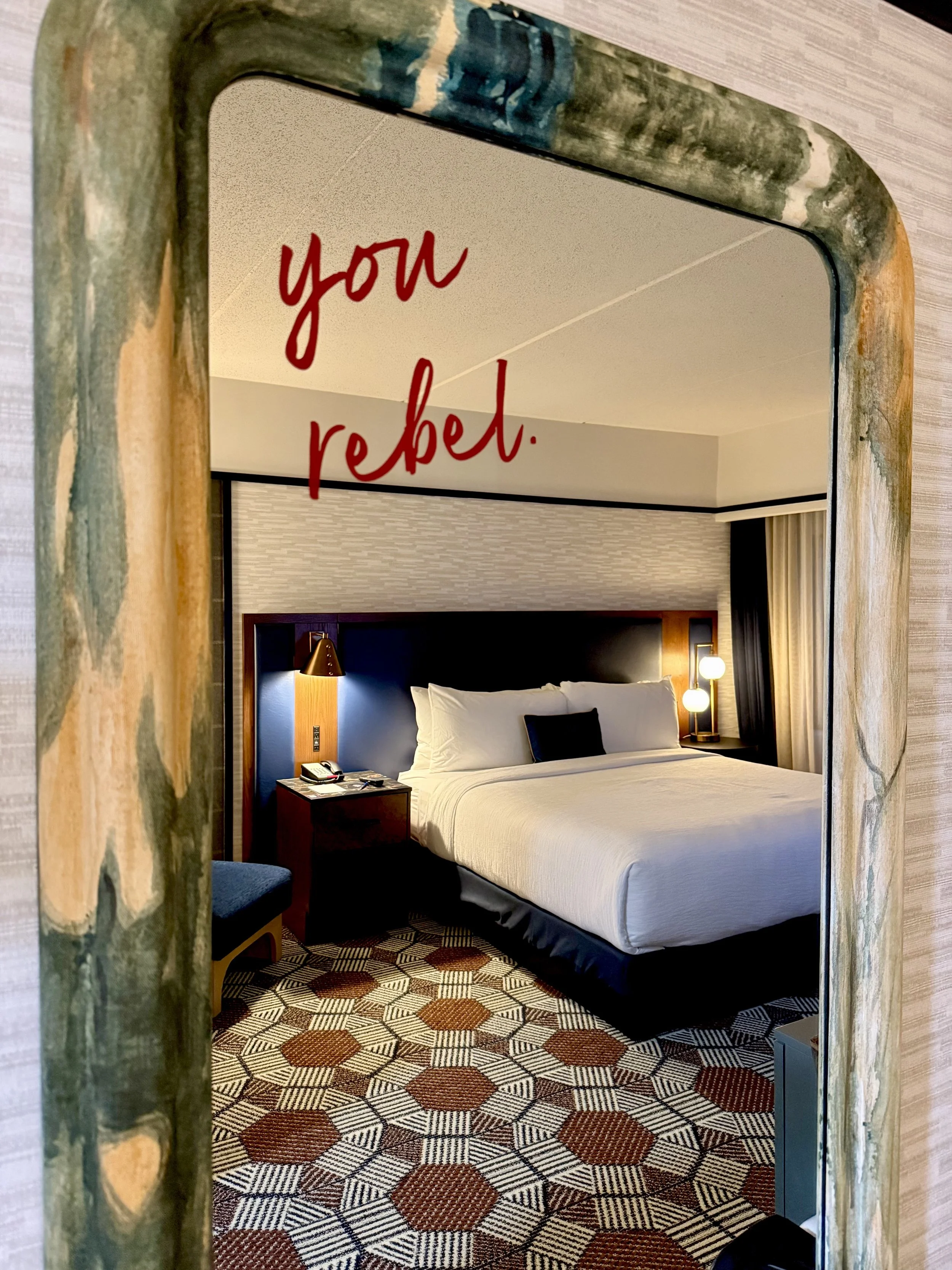 Guest room at The Mecklen Hotel in Charlotte reflected in a decorative mirror with the phrase “you rebel.” written in red script, featuring a king bed, modern lighting, and patterned flooring.