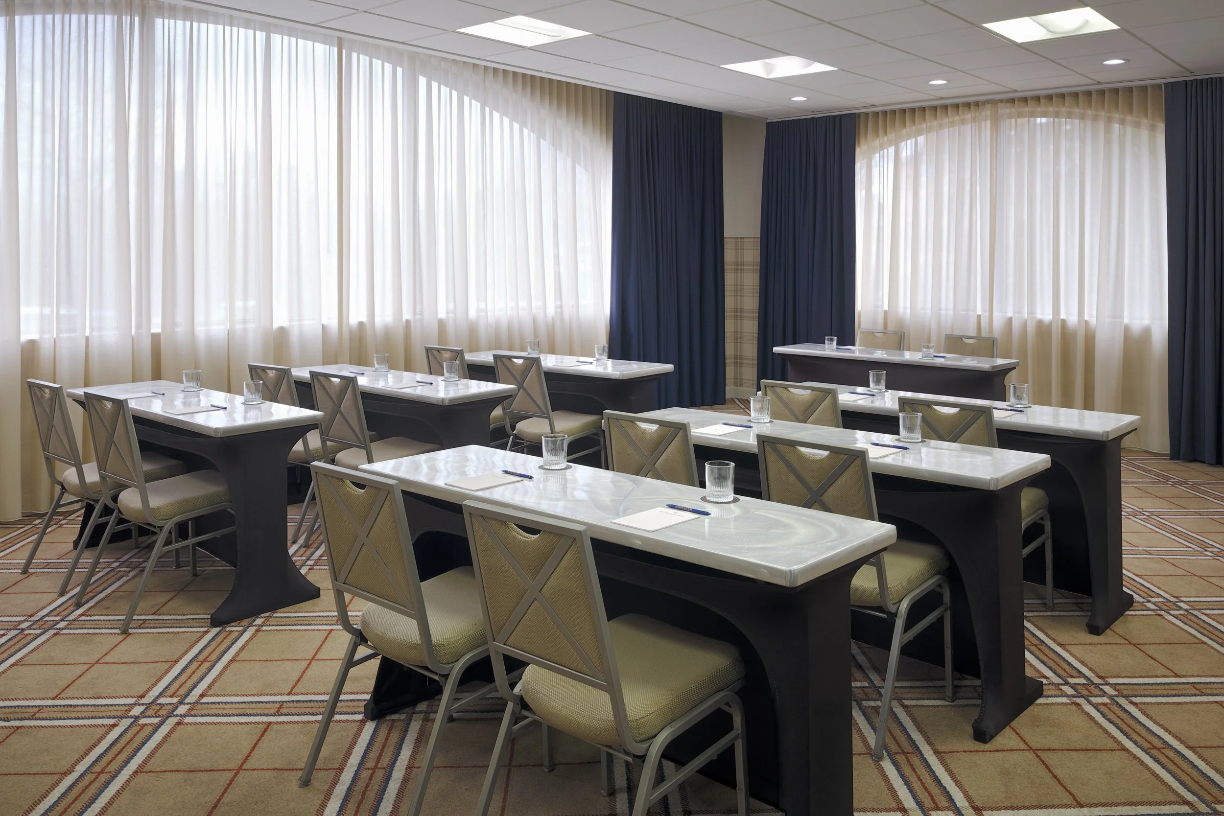 Meeting room at The Mecklen Hotel set in a classroom-style layout with tables, chairs, water glasses, and notepads, illuminated by natural light from large windows.