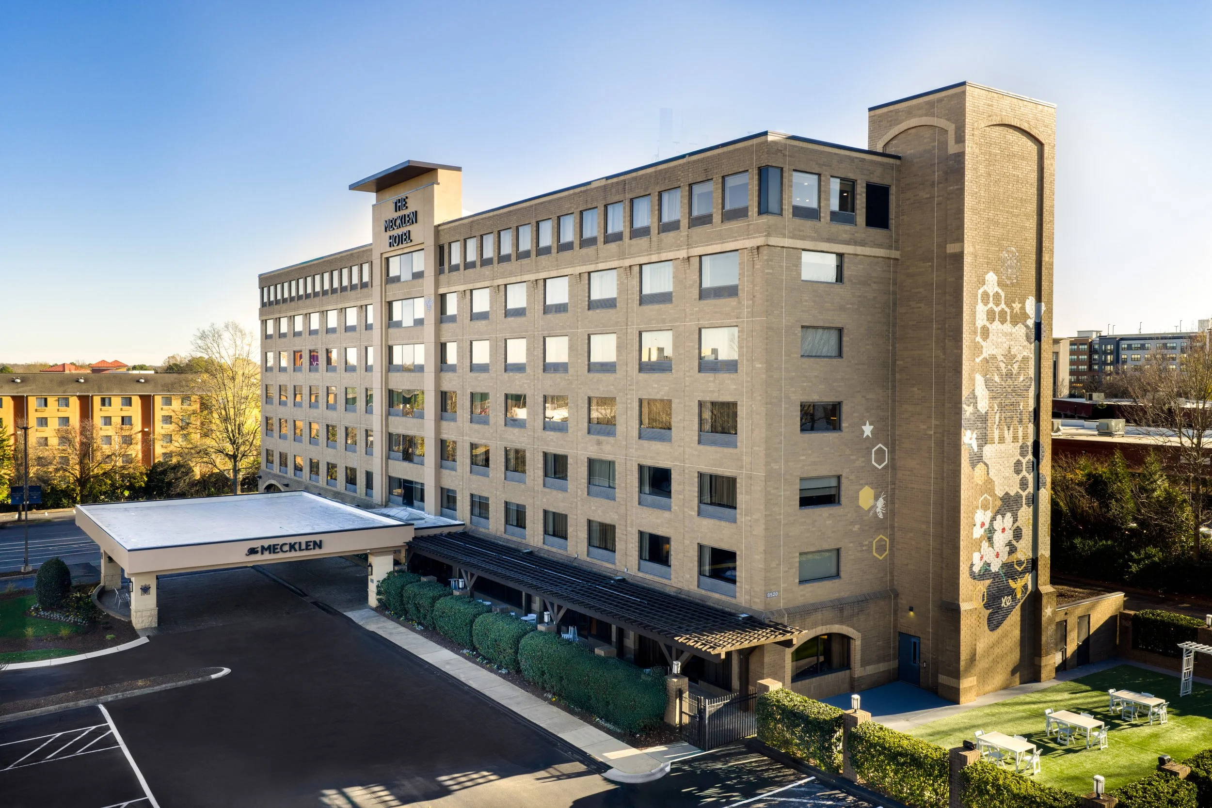 Daytime exterior of The Mecklen Hotel featuring a multi-story brick building, covered drive-up entrance, and landscaped outdoor area.