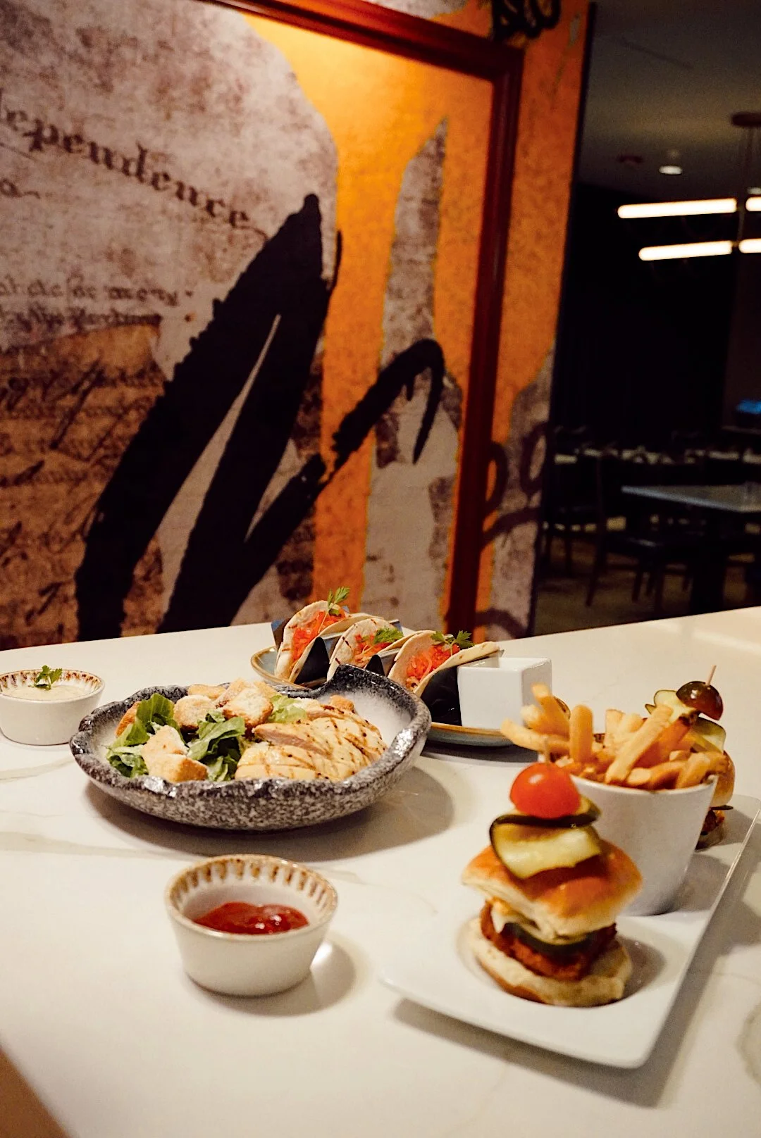 Grilled chicken Caesar salad, street tacos, and a crispy chicken slider with fries and pickles arranged on a bar counter in a modern restaurant setting.