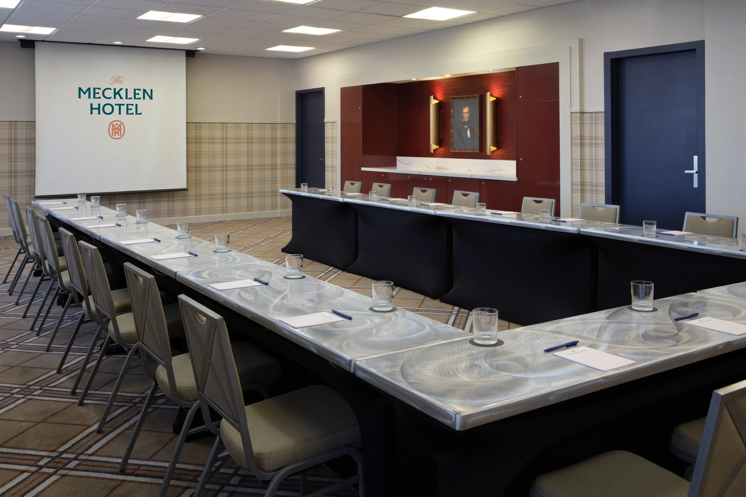 Conference room at The Mecklen Hotel set in a U-shape meeting layout with tables, chairs, water glasses, notepads, and a projection screen displaying The Mecklen Hotel logo.