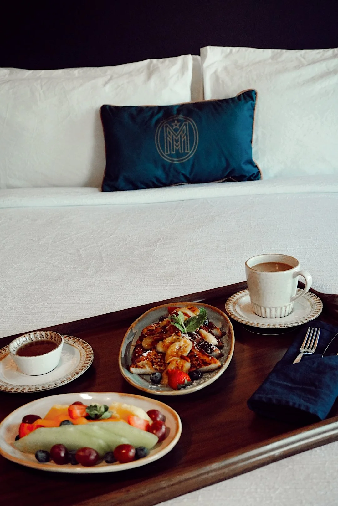 Breakfast in bed on a wooden tray with French toast topped with fruit, a side of fresh sliced fruit, a cup of coffee, and syrup, set against white bedding with a blue accent pillow.