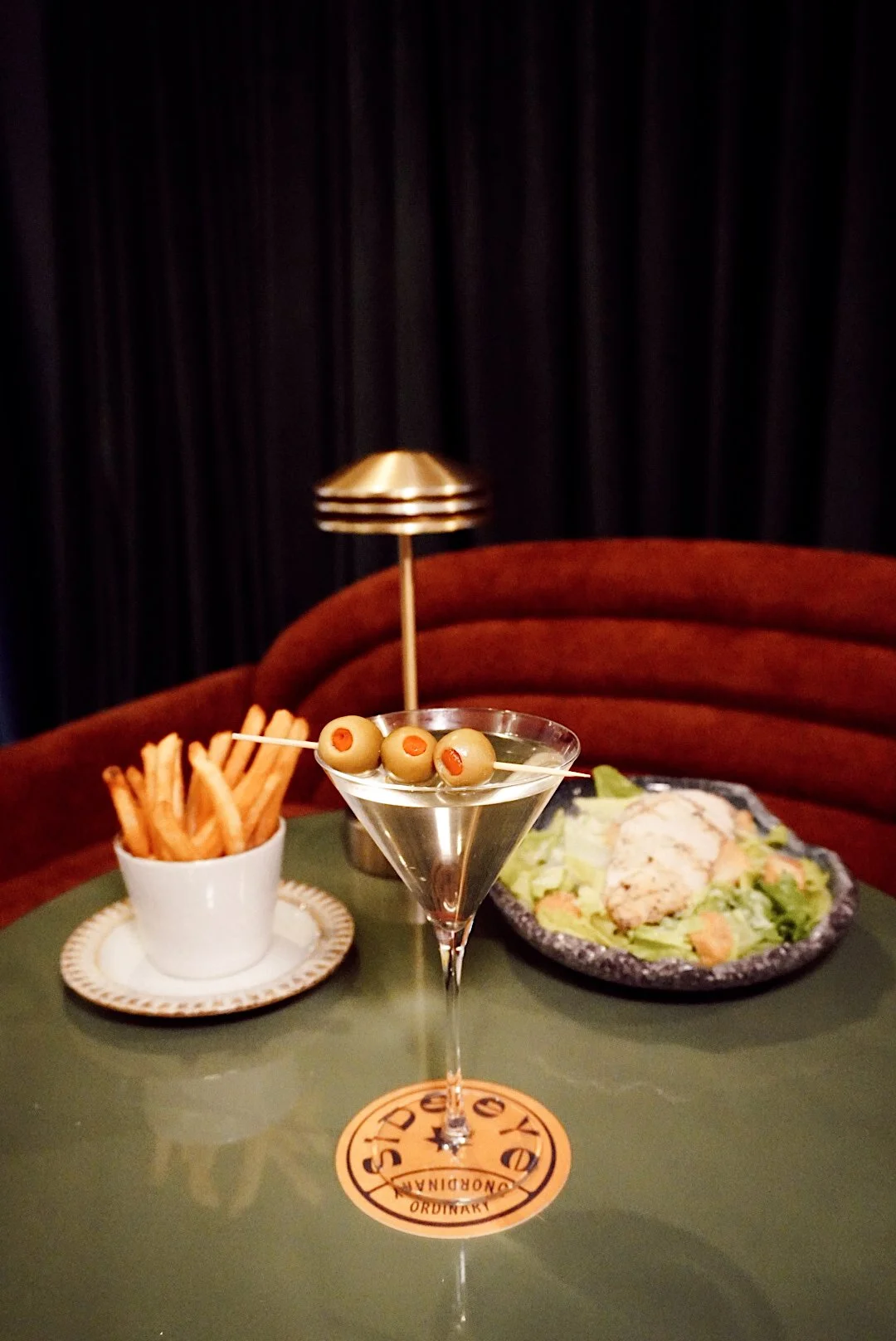 artini garnished with olives on a table with a side of fries and a chicken salad in a dimly lit lounge setting.