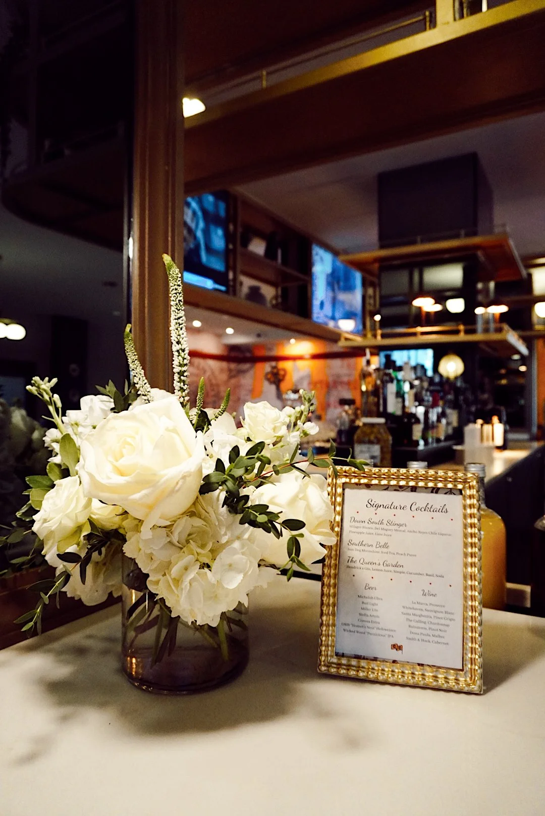 White floral arrangement and framed signature cocktail menu displayed on the bar at Side Eye restaurant inside The Mecklen Hotel.