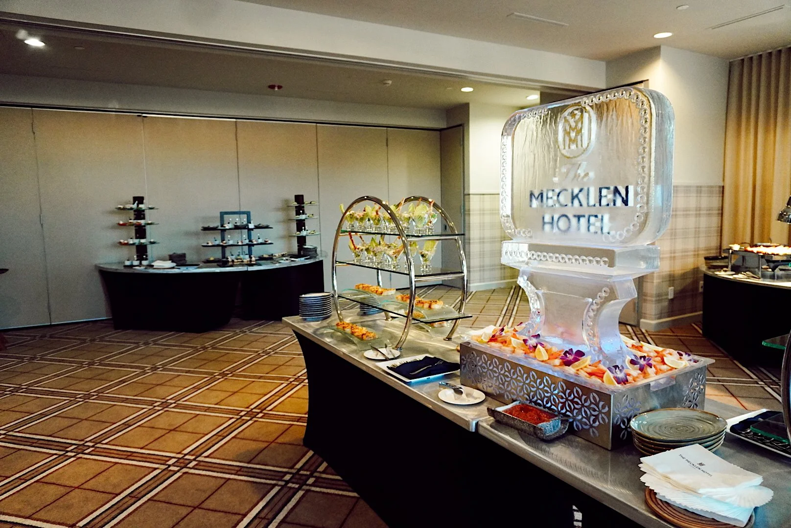 Catering buffet display at The Mecklen Hotel featuring appetizers, desserts, and an ice sculpture with The Mecklen Hotel logo in an event space.