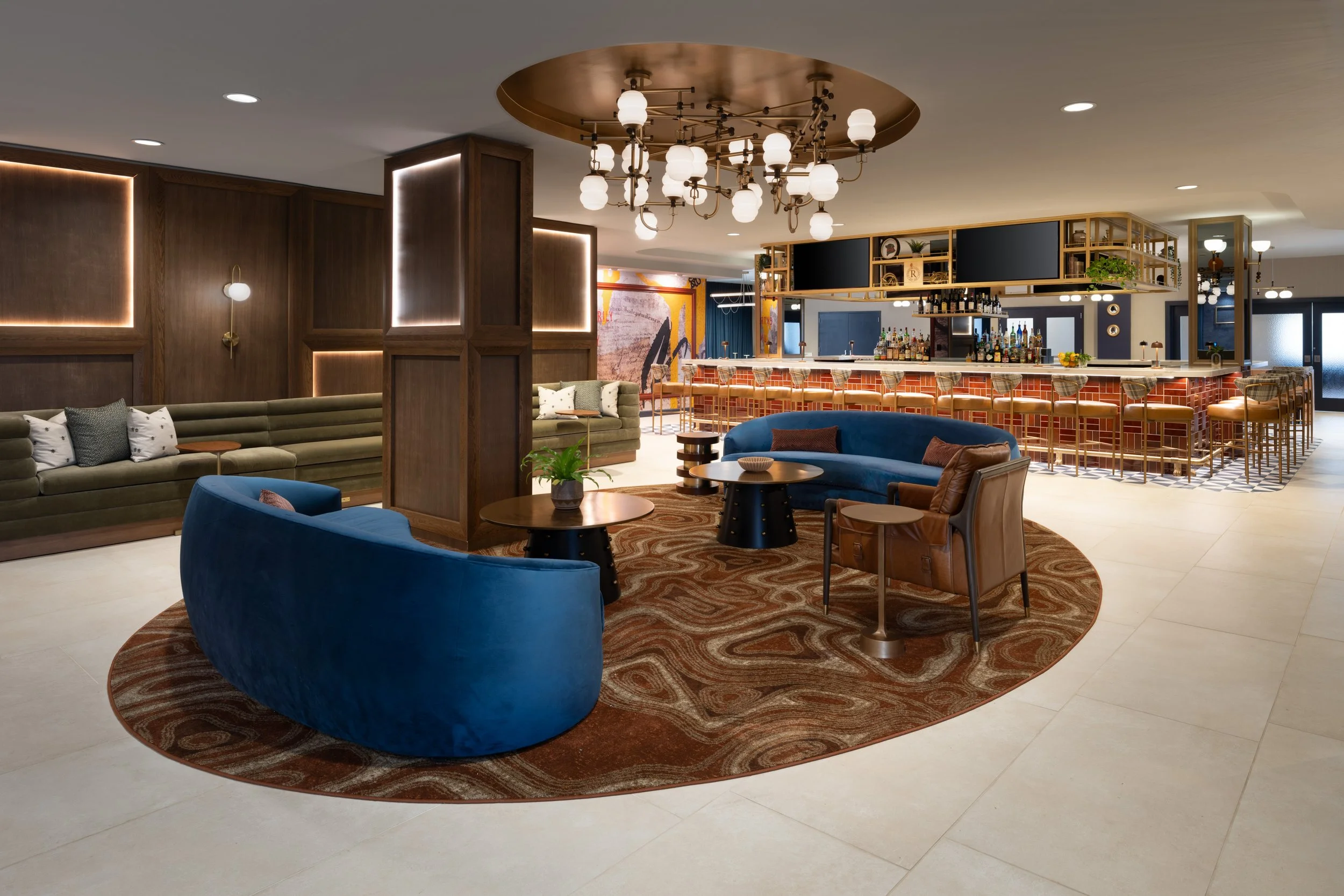 Stylish hotel lobby at The Mecklen featuring a circular seating area, and round tables on a patterned rug, set beneath a modern chandelier. In the background, the stylish Side Eye bar.