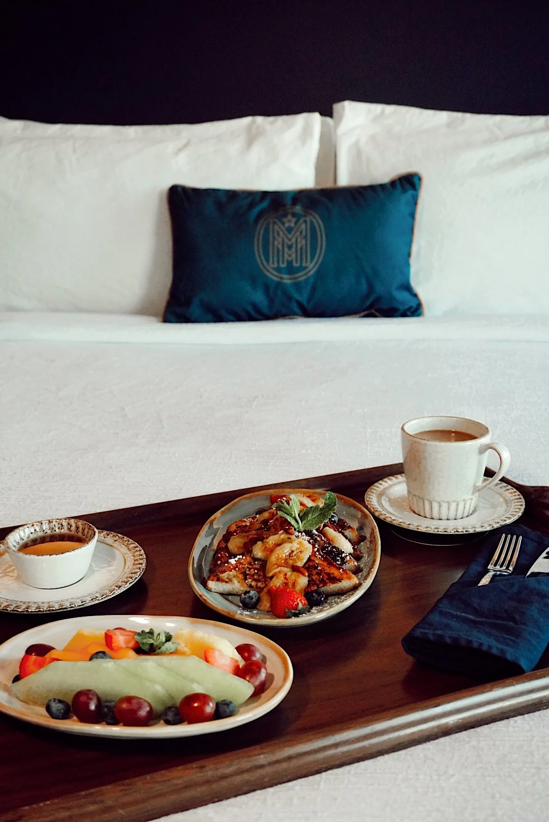 Breakfast tray with coffee, fresh fruit, and French toast served on a bed with The Mecklen Hotel branded pillow in the background.