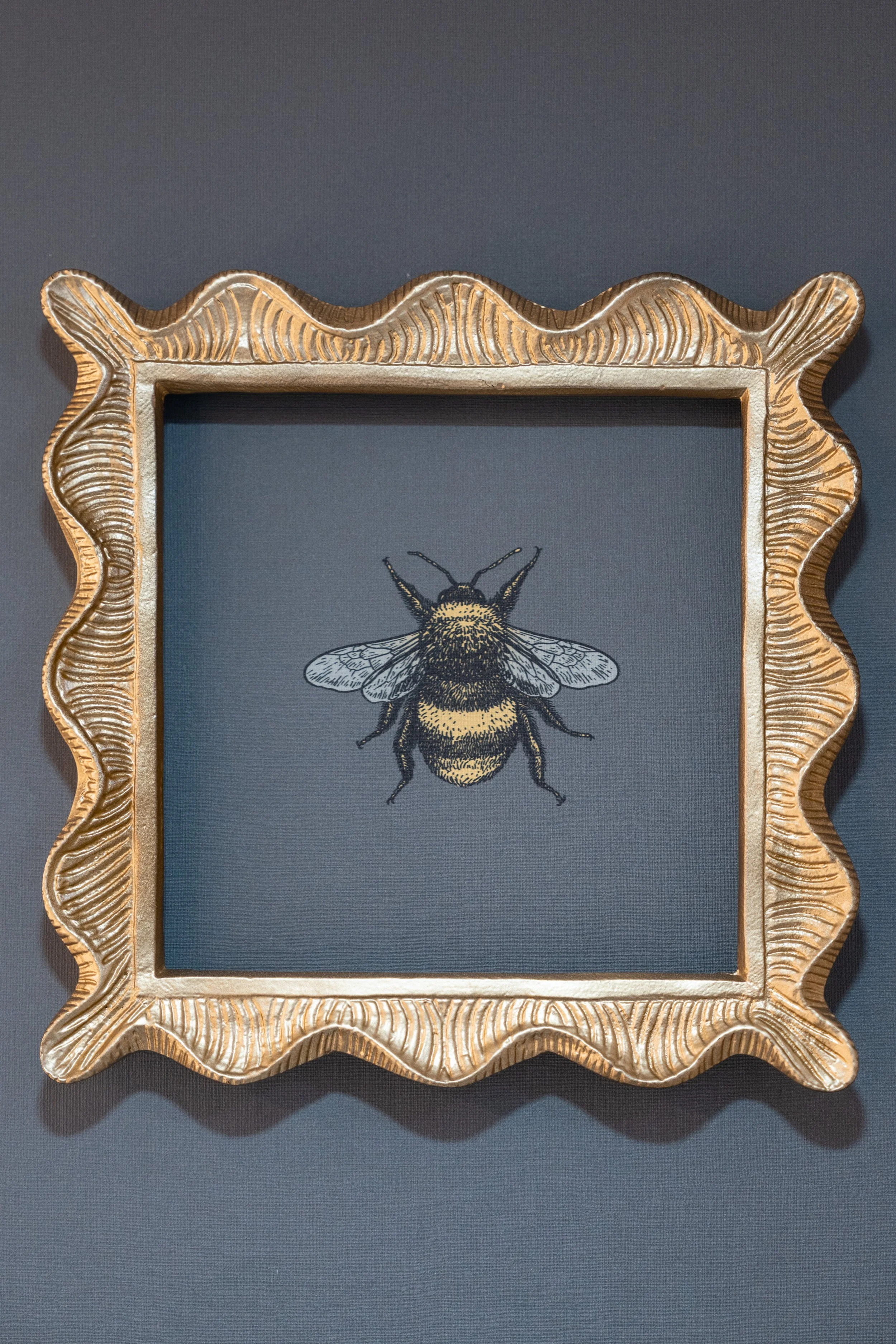 Framed illustration of a bumblebee on a dark background, set in an ornate gold wavy frame mounted on a wall.