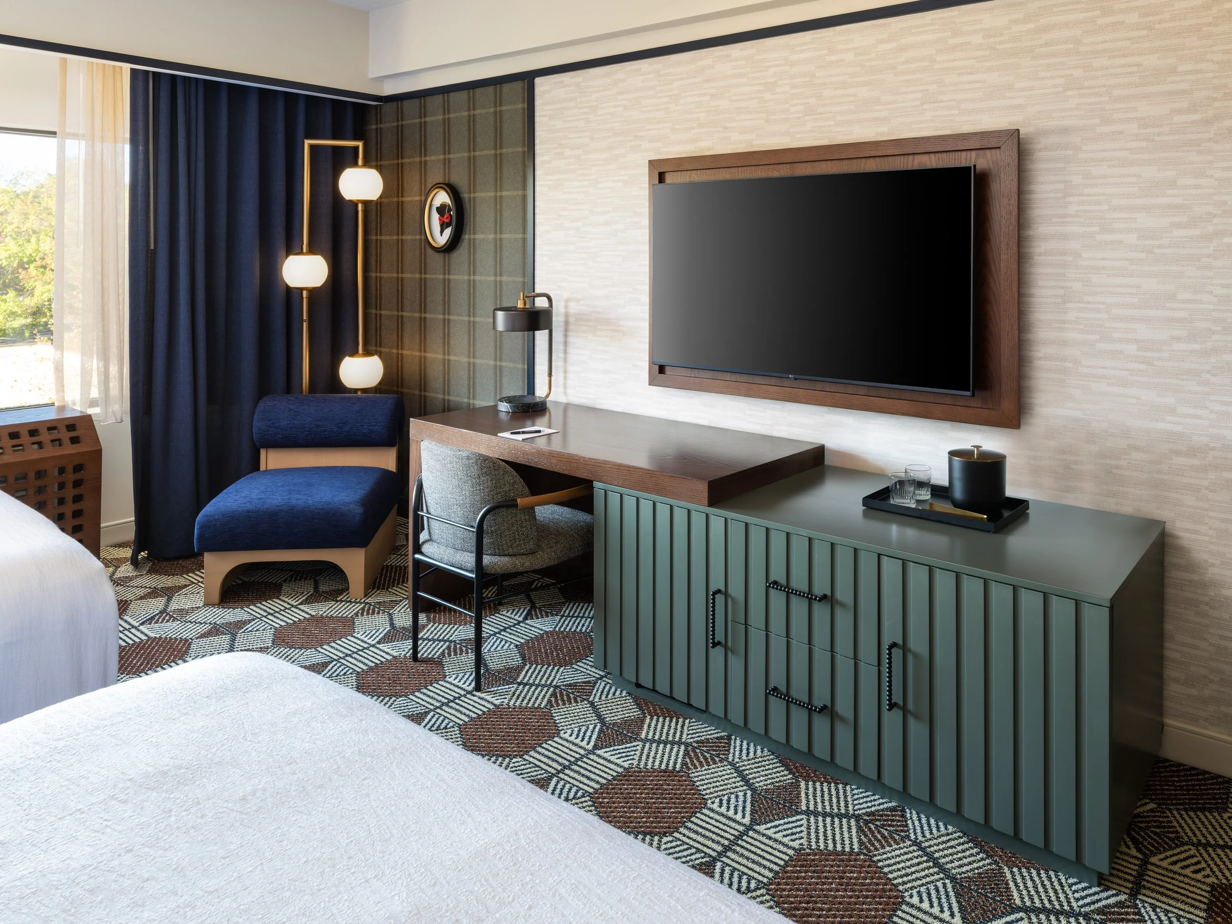 Desk space in Double Queen Room with a comfortable chair, lamp and television mounted above on the wall.