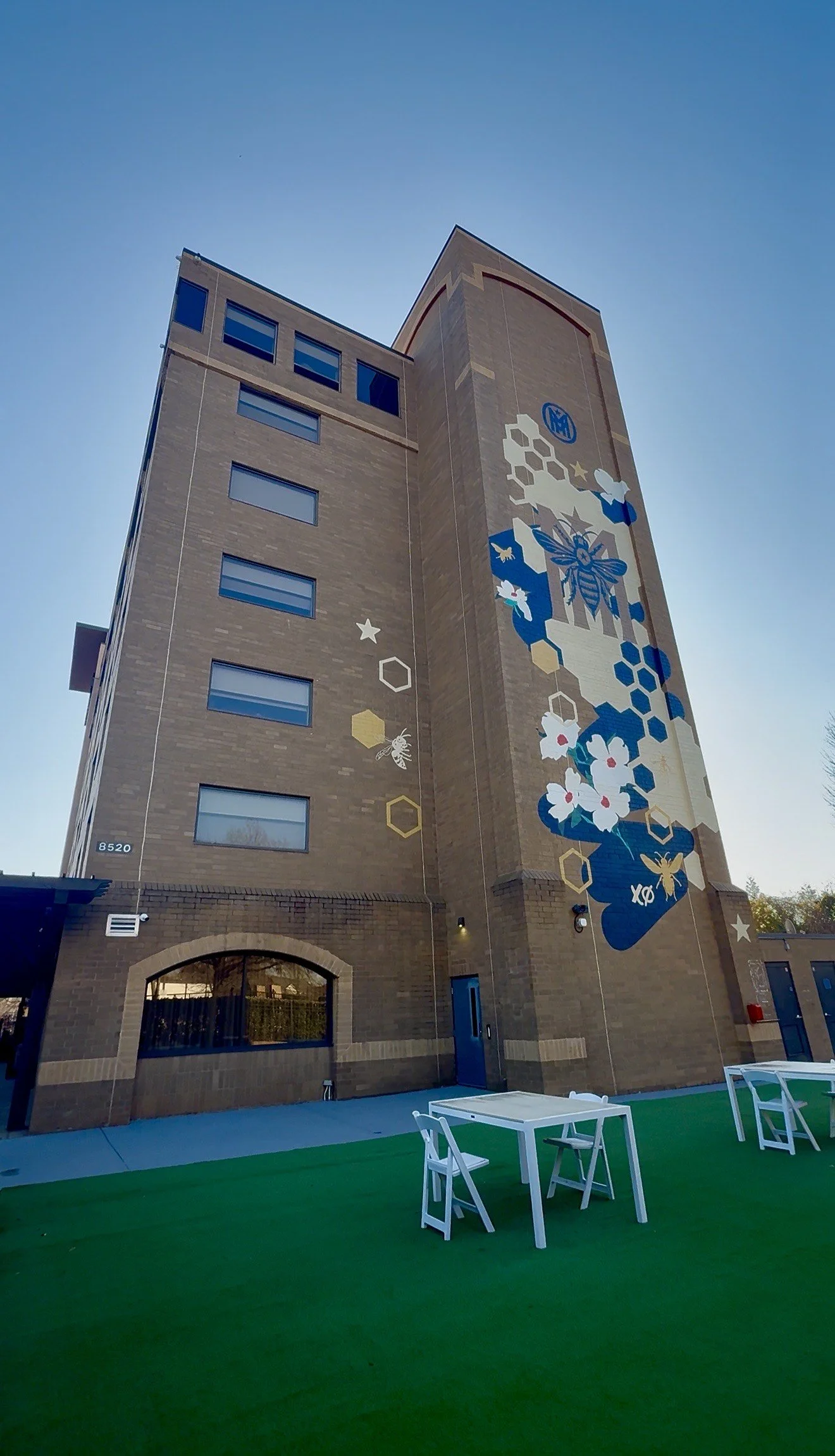 Exterior of The Mecklen Hotel in Charlotte featuring a large mural with bees, honeycomb patterns, and flowers on the brick façade, with outdoor seating on a green lawn patio.