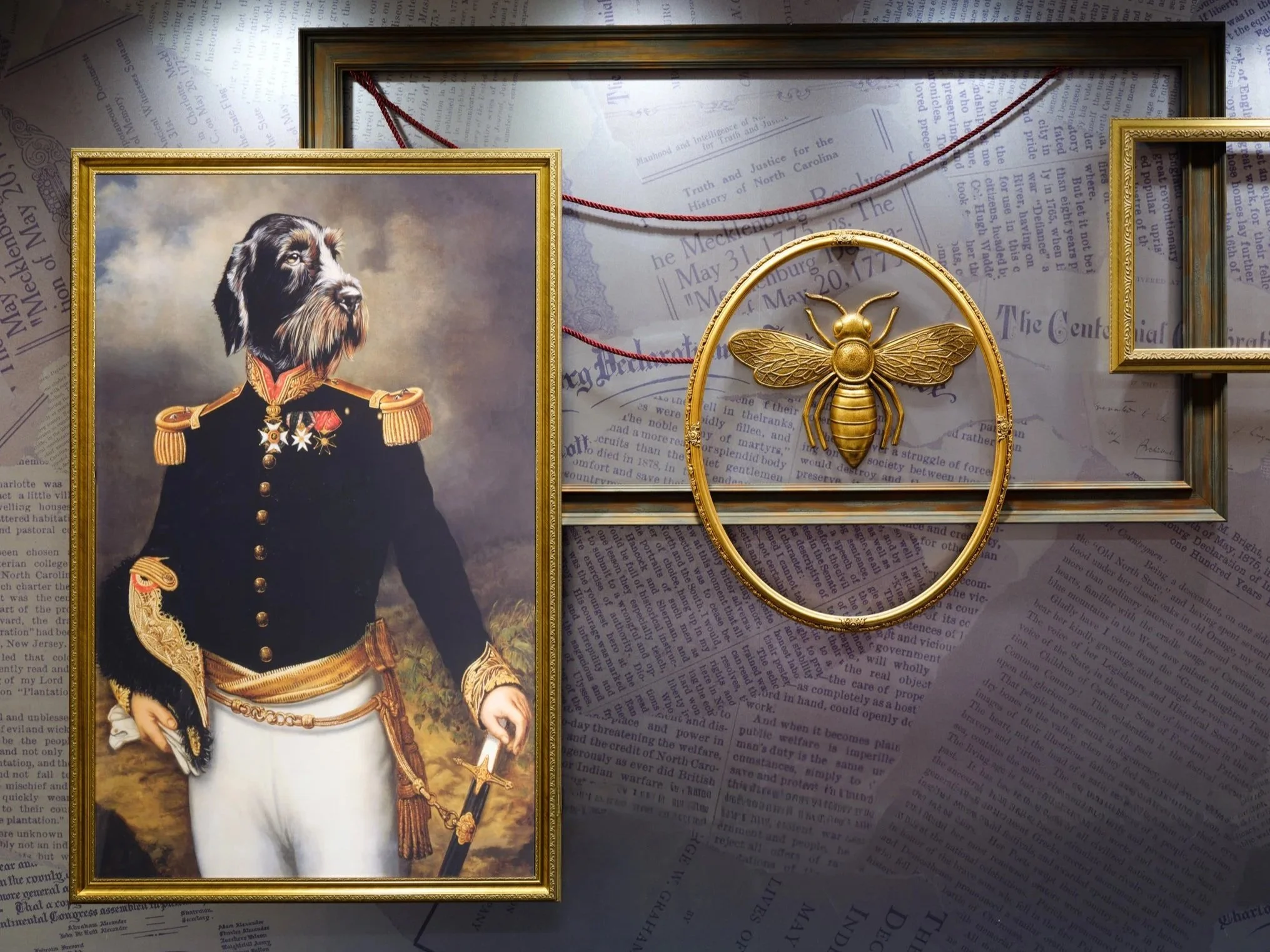Decorative wall display featuring vintage newspaper clippings, a gold bee emblem in a circular frame at center, and an empty frame to the right, alongside a portrait of a dog dressed in a formal military uniform on the left.