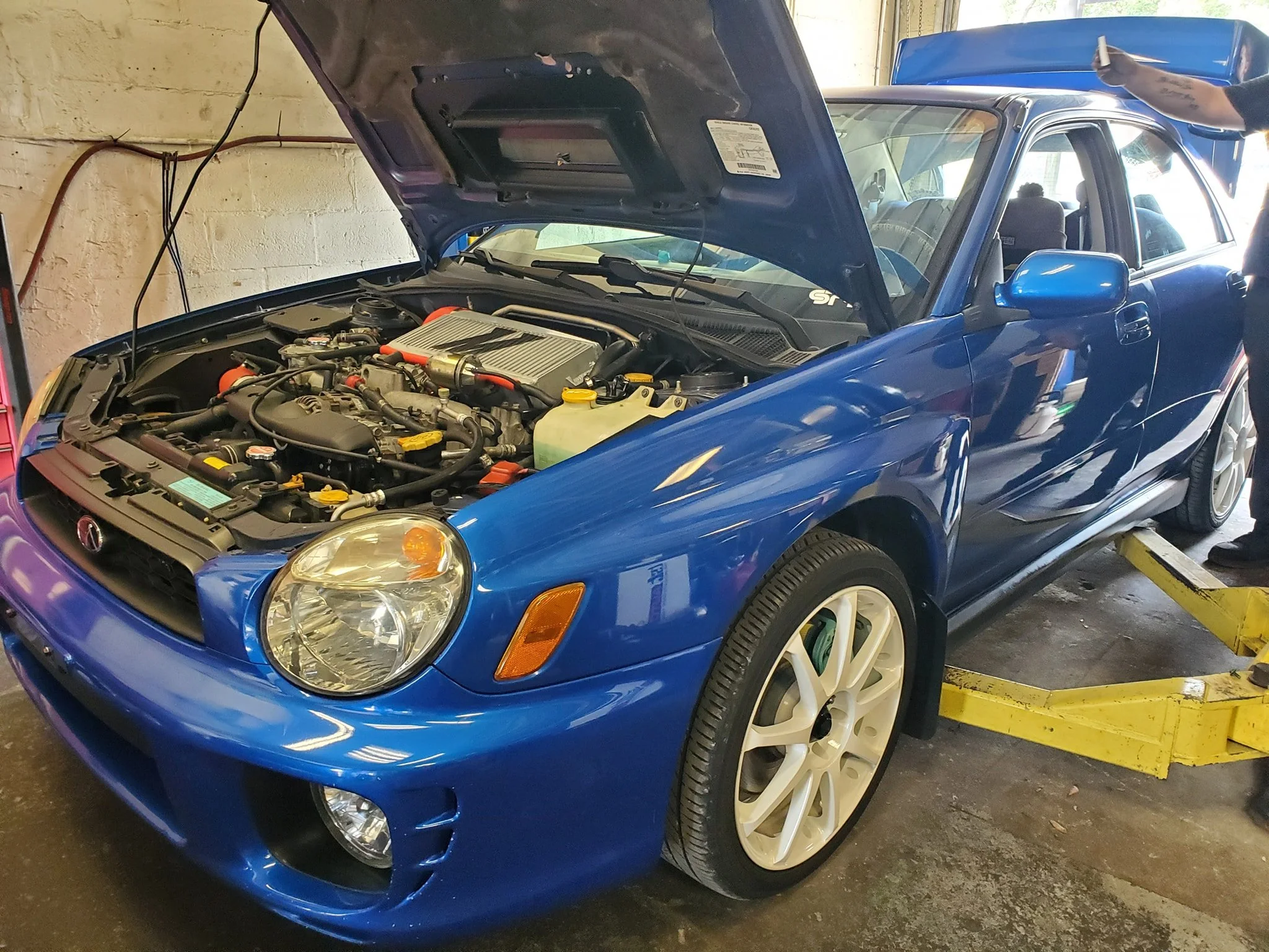 A blue Subaru sports car is being worked on in a garage with its hood open, revealing the engine.