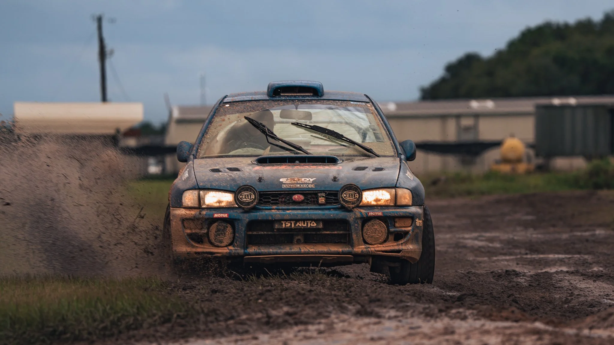 A blue rally car speeding through a muddy dirt track, kicking up dirt and mud in its wake, with a cloudy sky and industrial structures in the background.