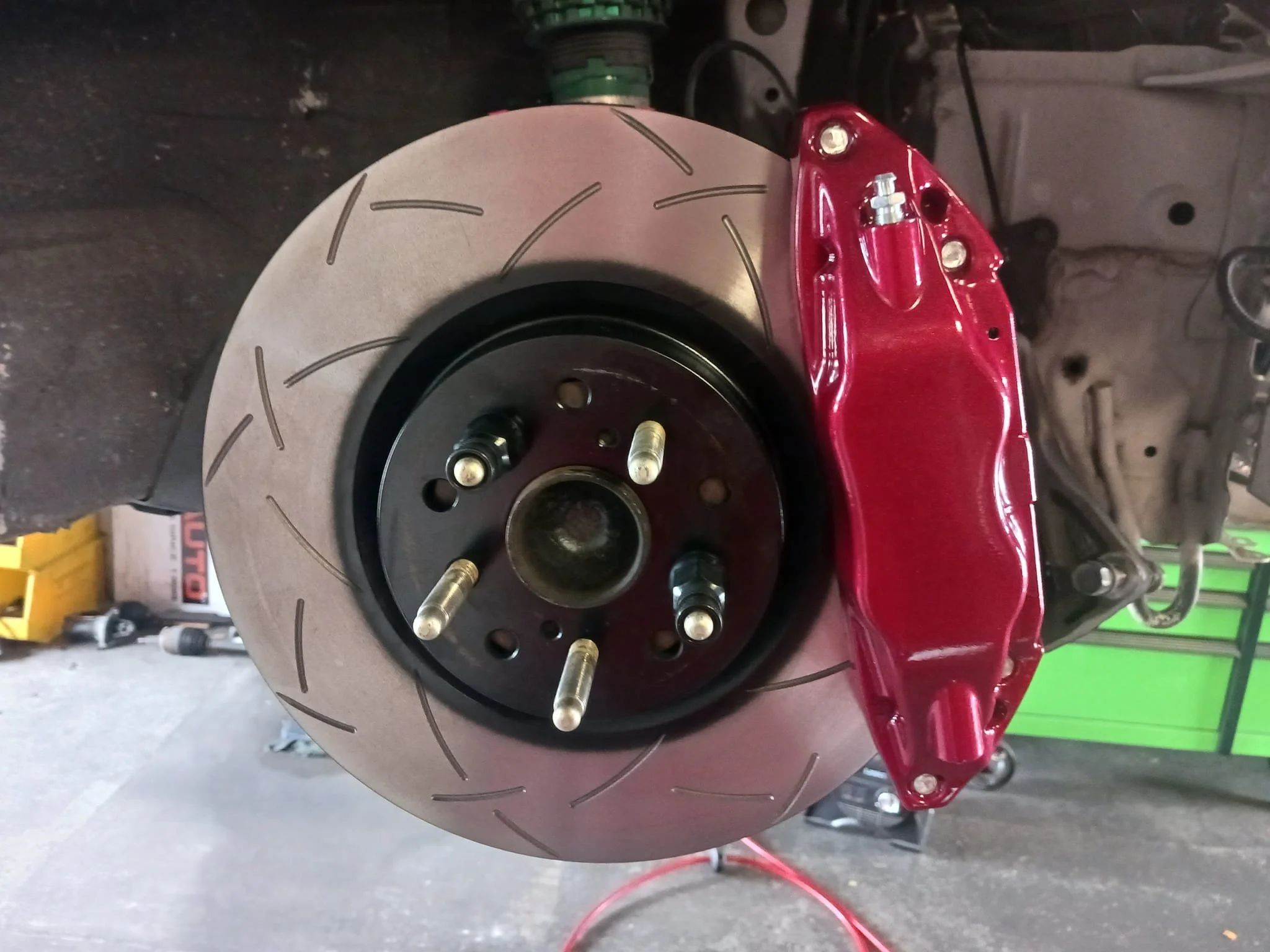 Close-up of a car's brake disc and red brake caliper, with wheel studs attached, in a garage setting.