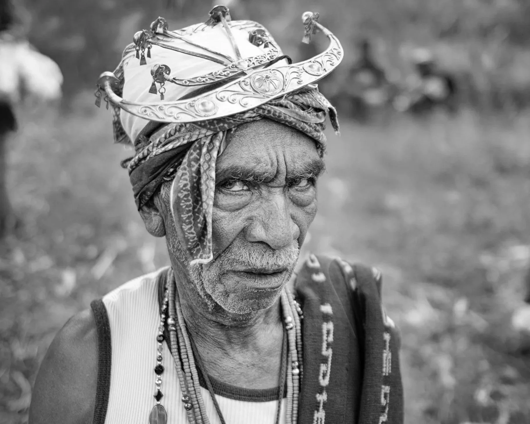 Black and white photo of an elderly man wearing traditional jewelry, a headscarf, and a vest. The man has a serious expression and is looking slightly to the side, with a blurred outdoor background.