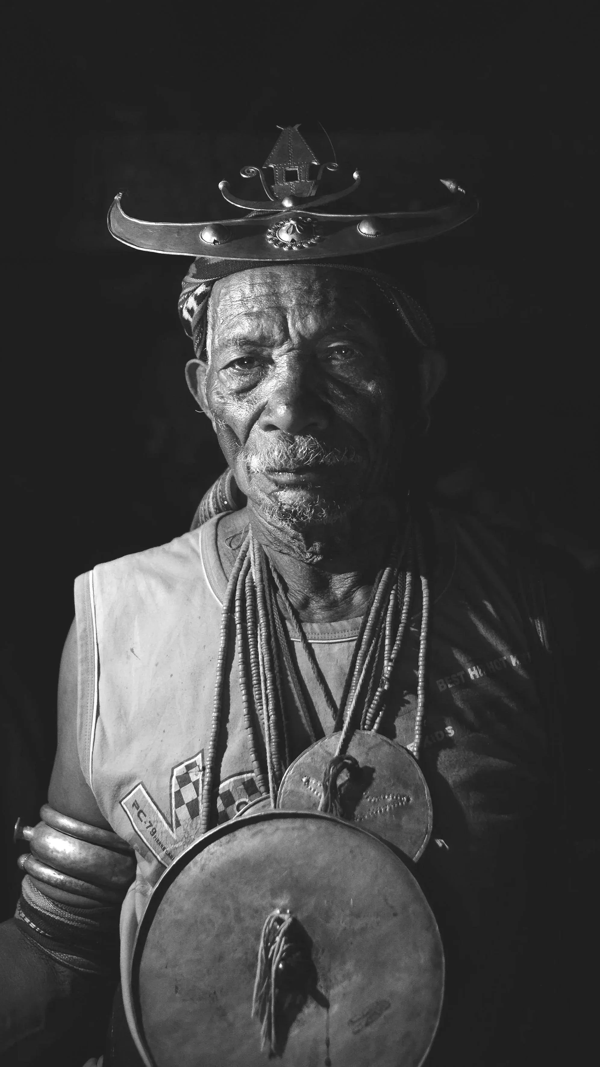 A black and white photo of an elderly man with a serious expression, wearing a striped bandana on his head, multiple necklaces, and a tank top, with a large medallion hanging from his neck. He is holding a drum, with a fish hanging from a string over his chest, and an ornate metal crown balanced on his head.